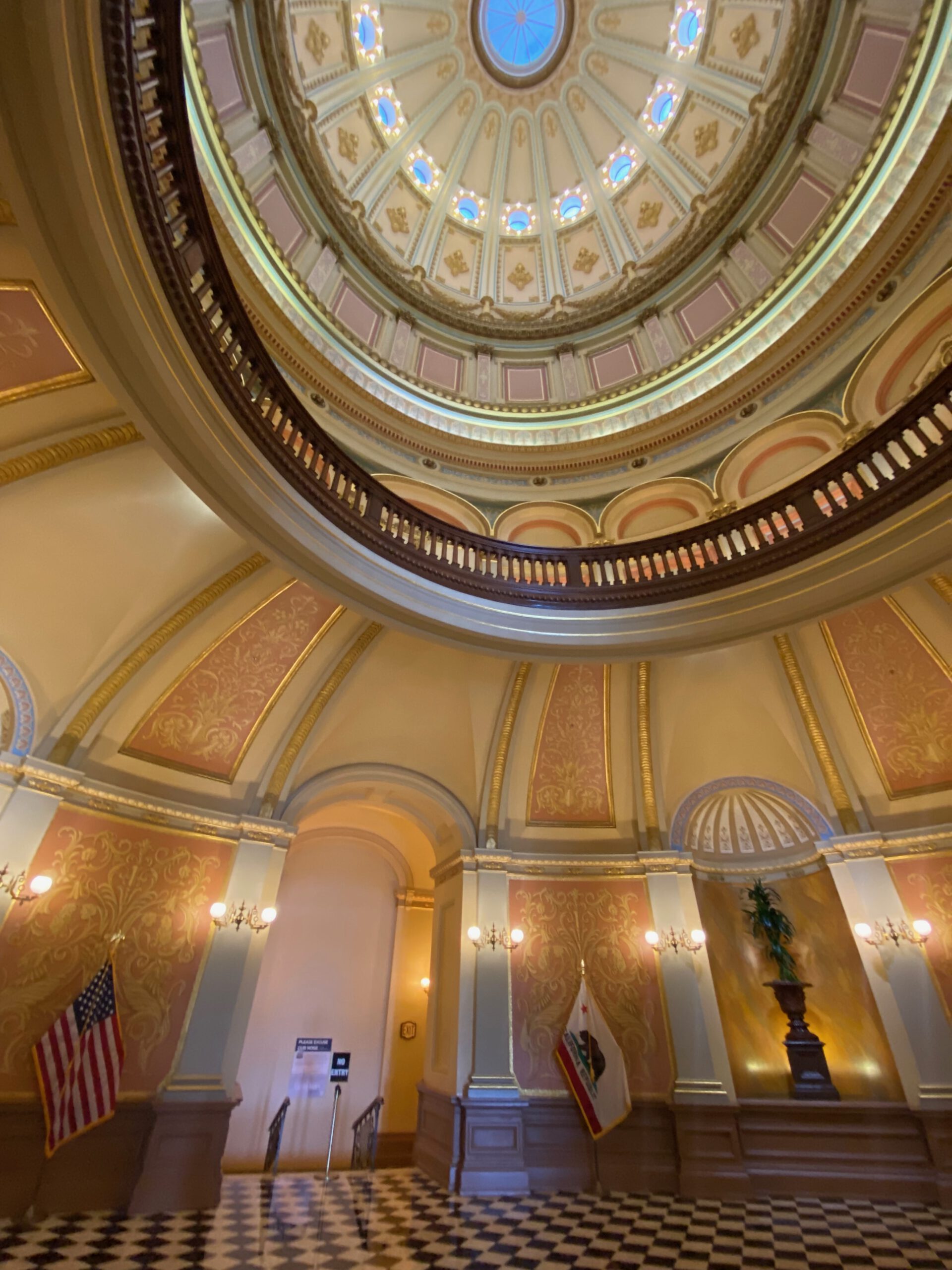Central rotunda inside the California State Capitol Museum, featuring the ornate historic dome with decorative panels, circular balcony, American and California flags, and a black and white marble floor beneath the skylight.