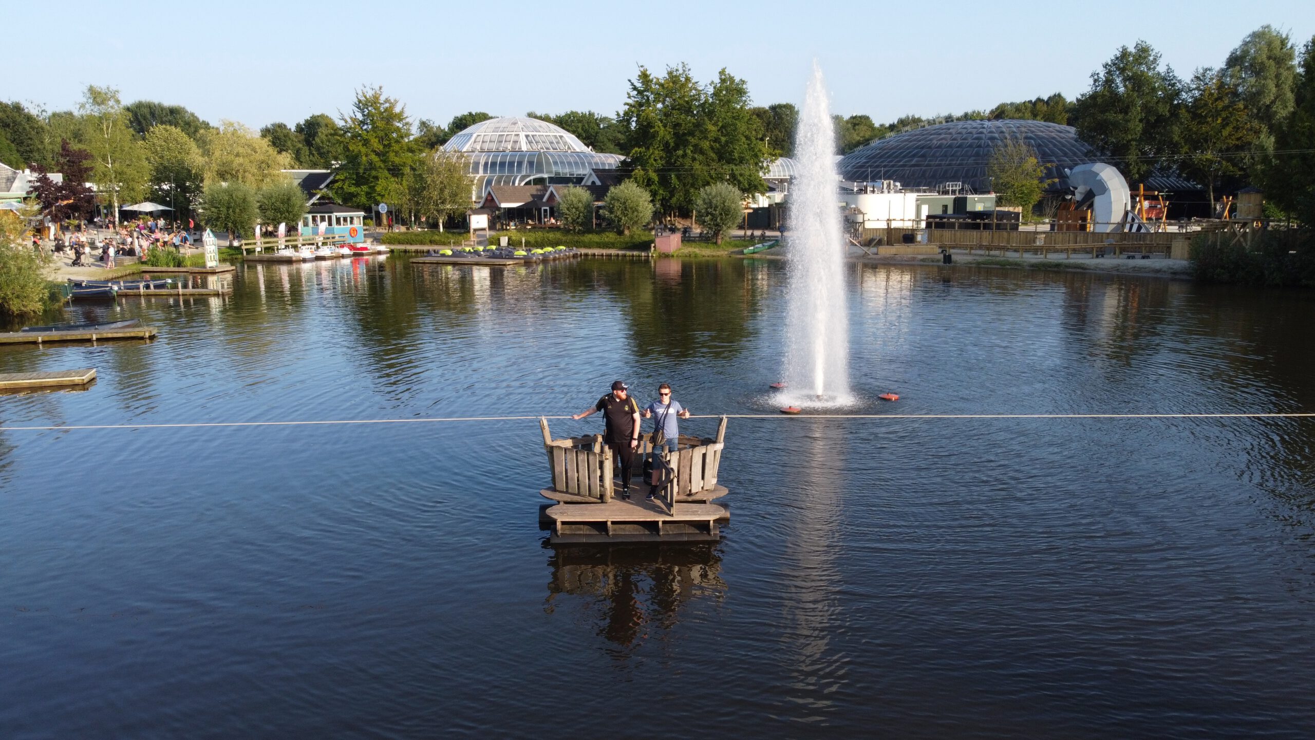 Center Parcs De Huttenheugte Day Visit drone shot above the central lake, Peter Smit and Timeuw Timo van den Berg standing on a small wooden raft platform with water fountain and the Aqua Mundo domes of Center Parcs De Huttenheugte in the background in Drenthe.