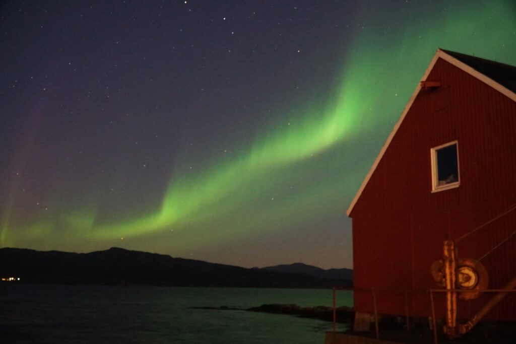 Hjertholmen Lødingen Northern Lights arching over calm Arctic waters beside a red wooden building