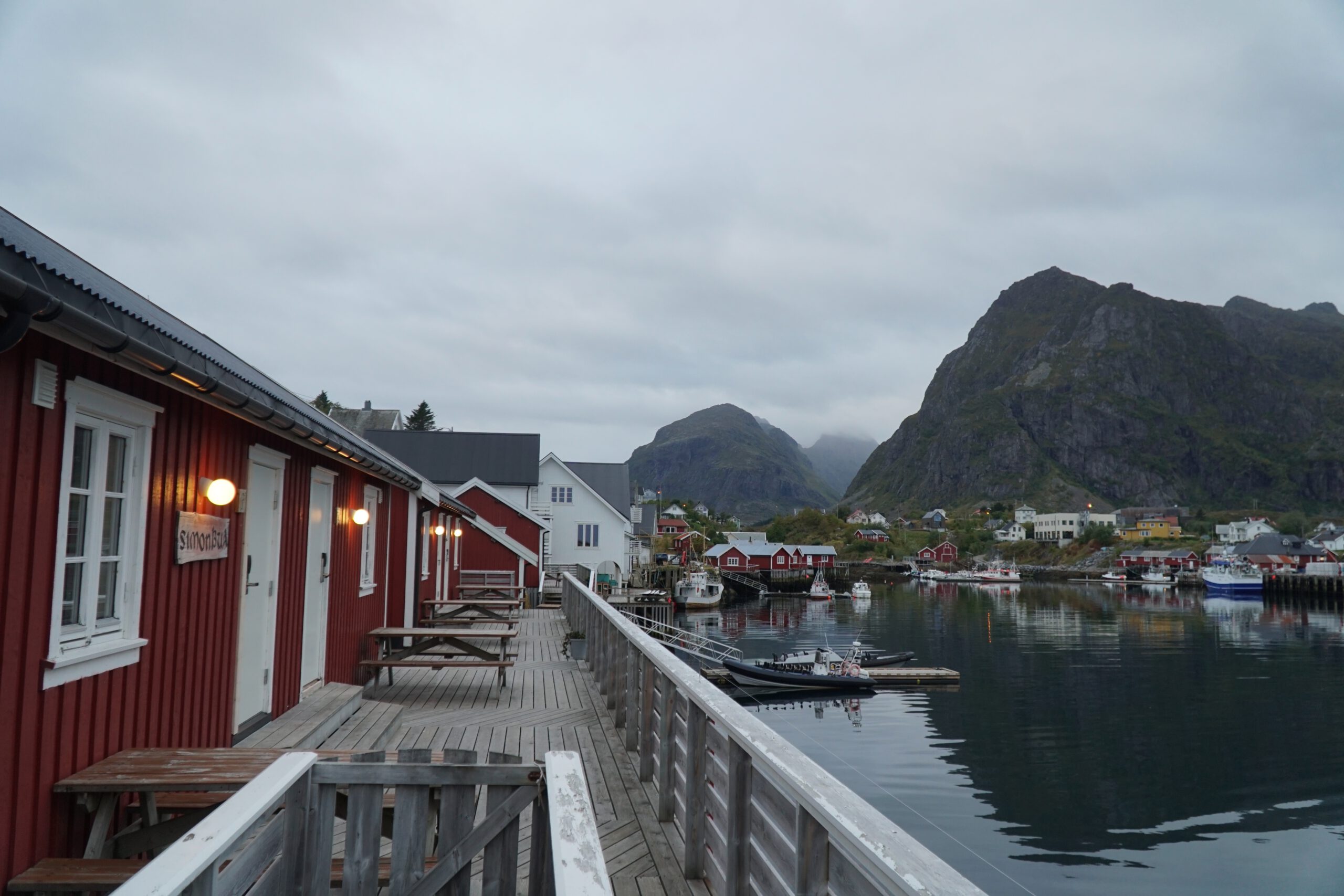 The Tide Fisherman Cabins Sørvågen wooden boardwalk with red fisherman cabins, small boats in the harbor and mountains surrounding Sørvågen village