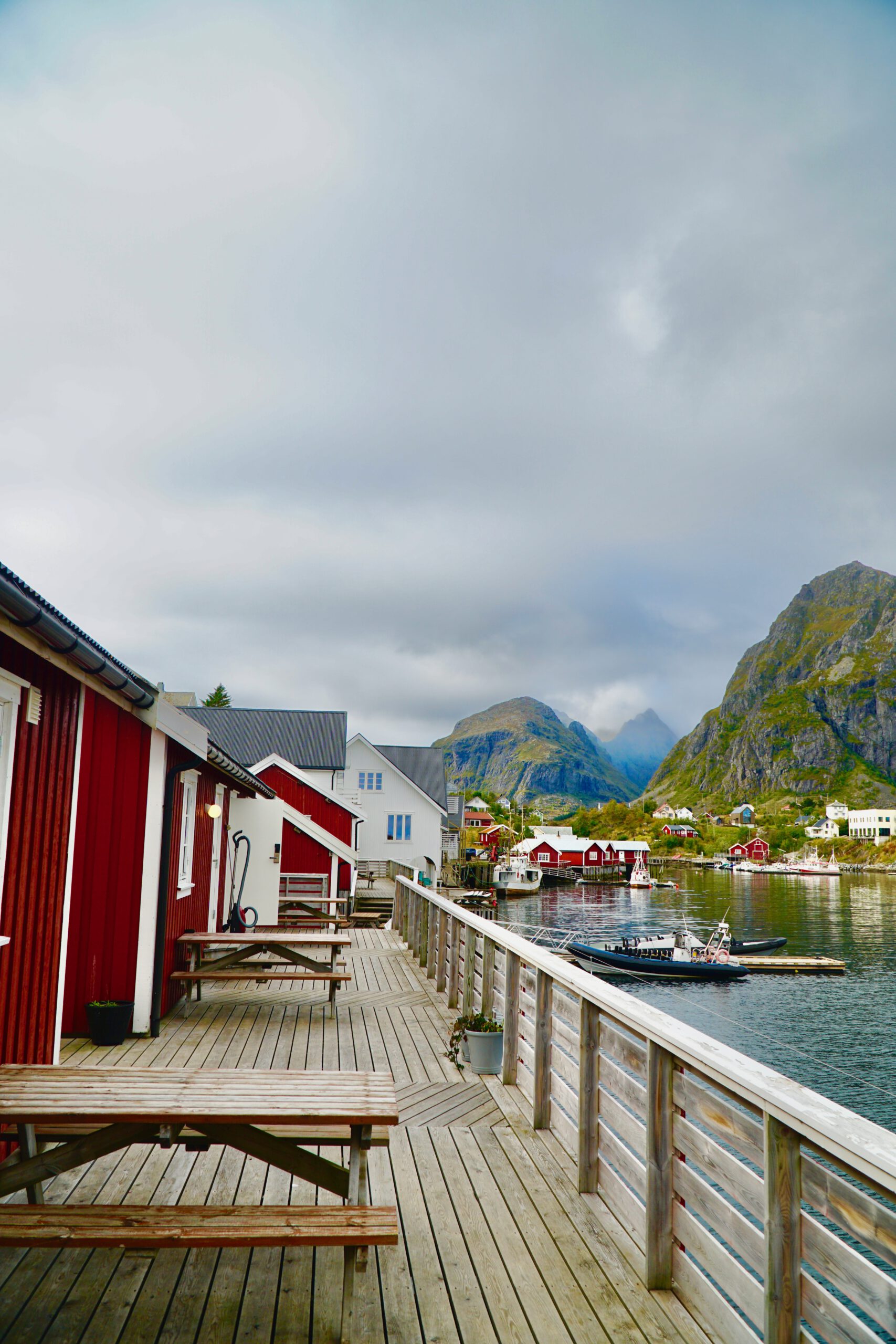 The Tide Fisherman Cabins Sørvågen wooden boardwalk with red rorbuer cabins, harbor water, boats and dramatic Lofoten mountains in the background