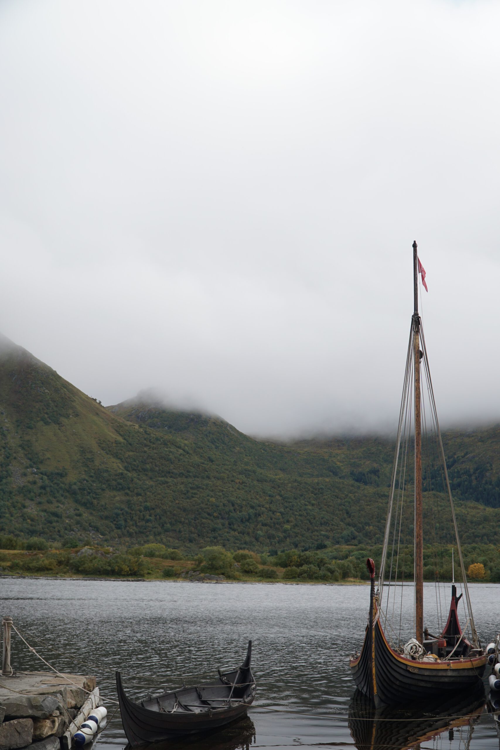 Lofotr Viking Museum outdoor Viking harbor with traditional Viking ships moored by the water, surrounded by misty mountains at the Borg site on Vestvågøy