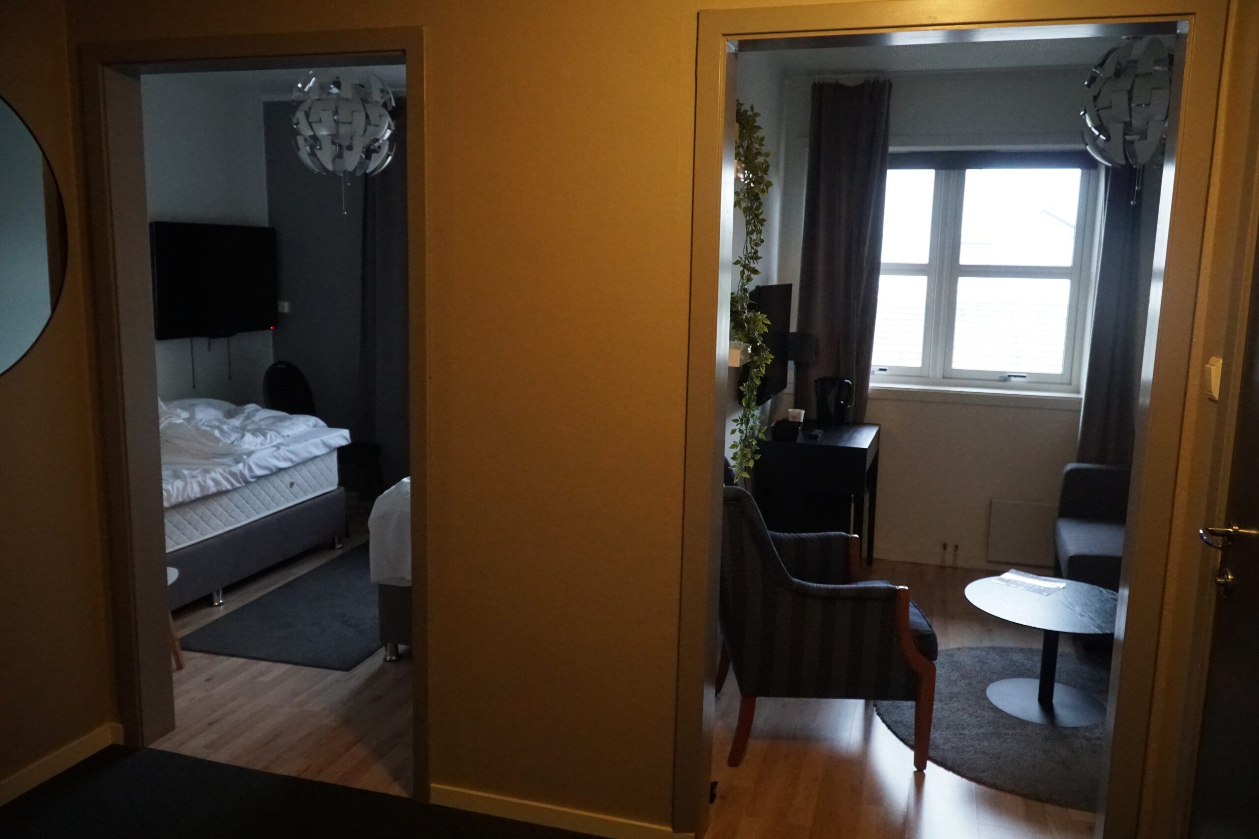 Interior view of Mosjøen Romutleie Sentrum 3 showing the bedroom with two single boxspring beds and a separate living area with sofa, chair, table, and window, a practical budget room layout in Mosjøen, Norway