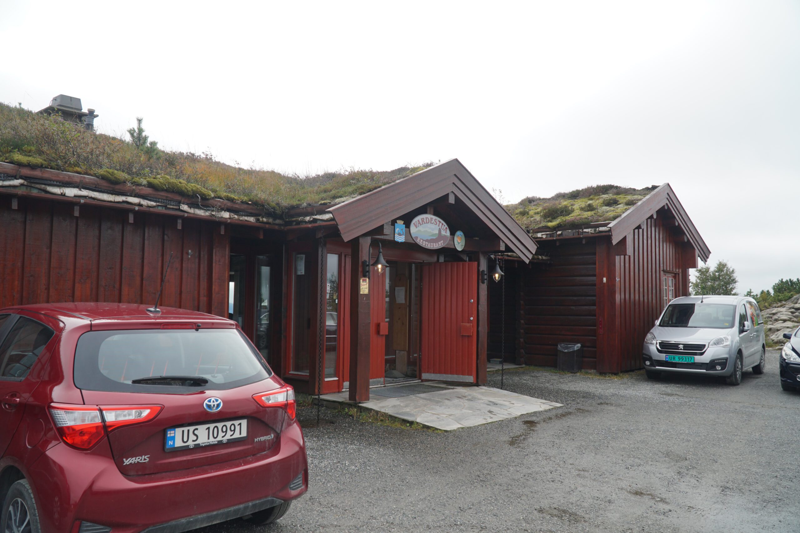 Vardestua Restaurant Molde view entrance at Varden, traditional red wooden building with grass roof at 407 meters above sea level, cars parked outside in Molde Norway under cloudy sky.