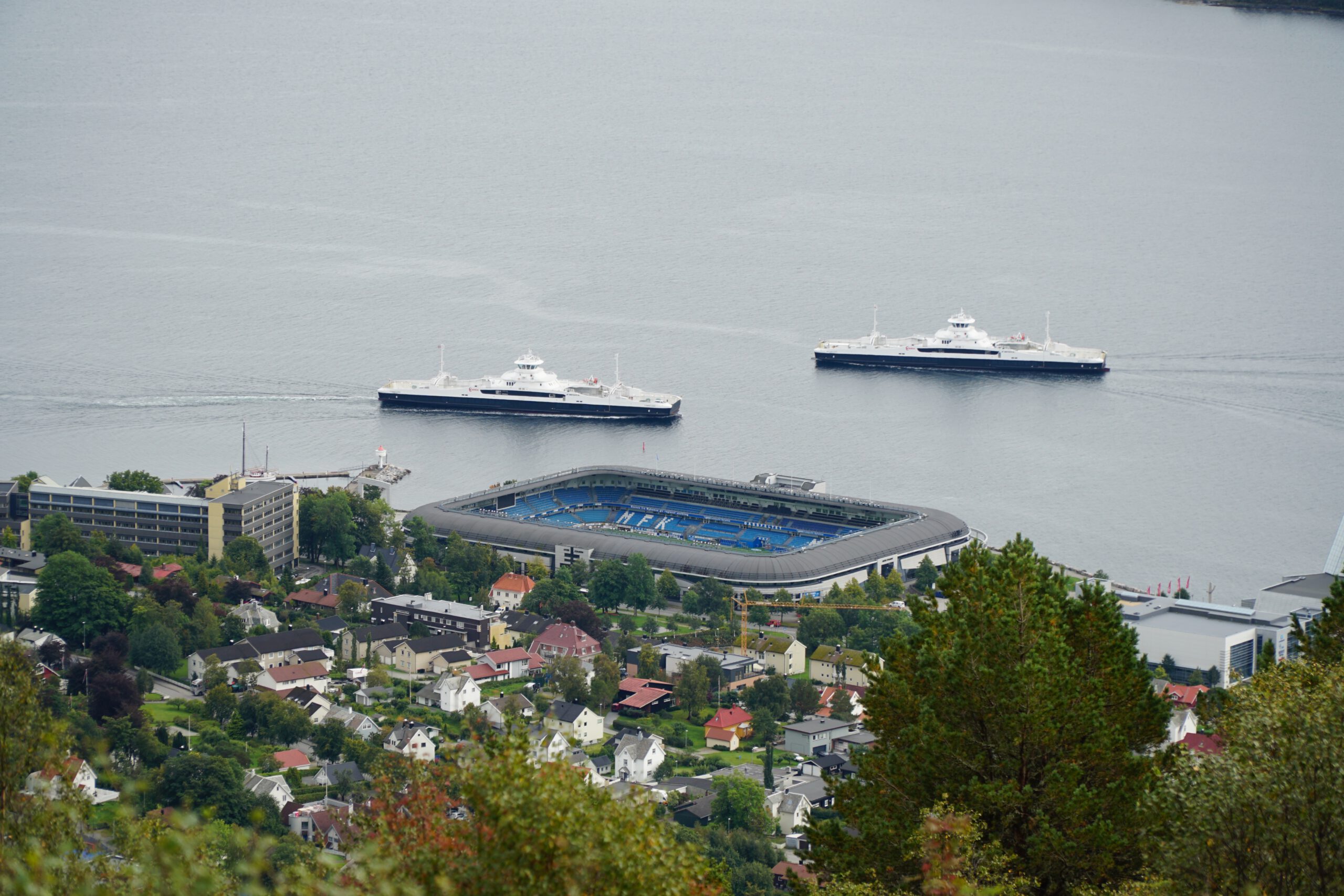 Aker Stadion Molde viewed from Vardestua panoramic viewpoint, the Molde FK stadium located directly along the Romsdalsfjord with two ferries sailing behind the waterfront arena and residential houses in the foreground.