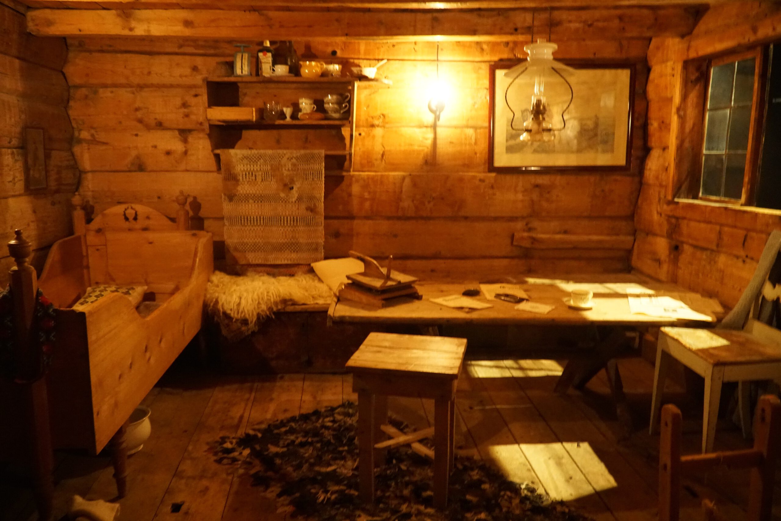 Traditional wooden farmhouse interior at Norsk Fjordsenter Geiranger featuring rustic timber walls, carved bed, oil lamp, writing desk with papers and coffee cup, part of the UNESCO World Heritage Geirangerfjord cultural exhibition