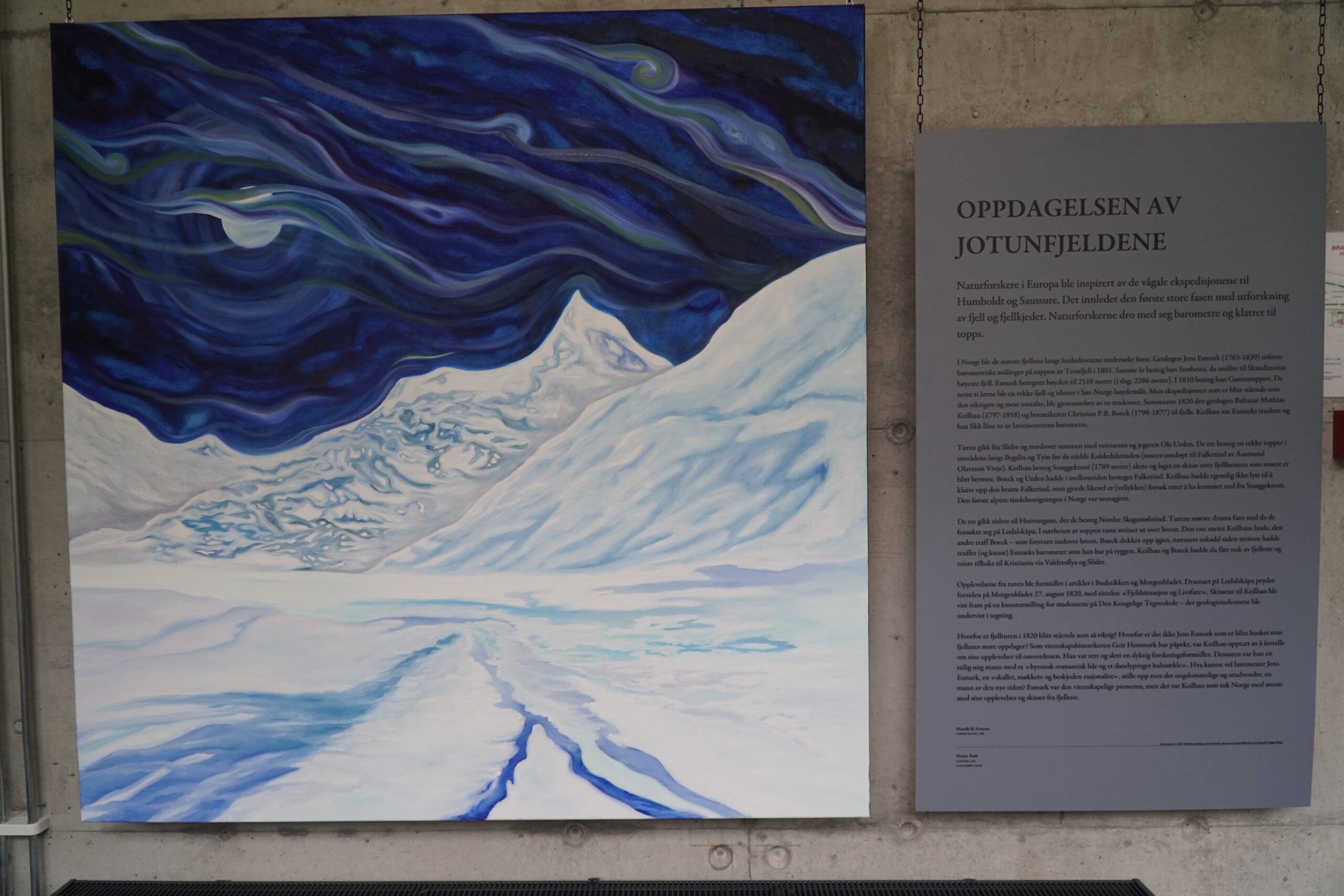 Large blue glacier painting inside Norsk Fjordsenter Geiranger showing swirling northern sky above a snow covered mountain landscape, with information panel about the discovery of Jotunheimen on concrete wall