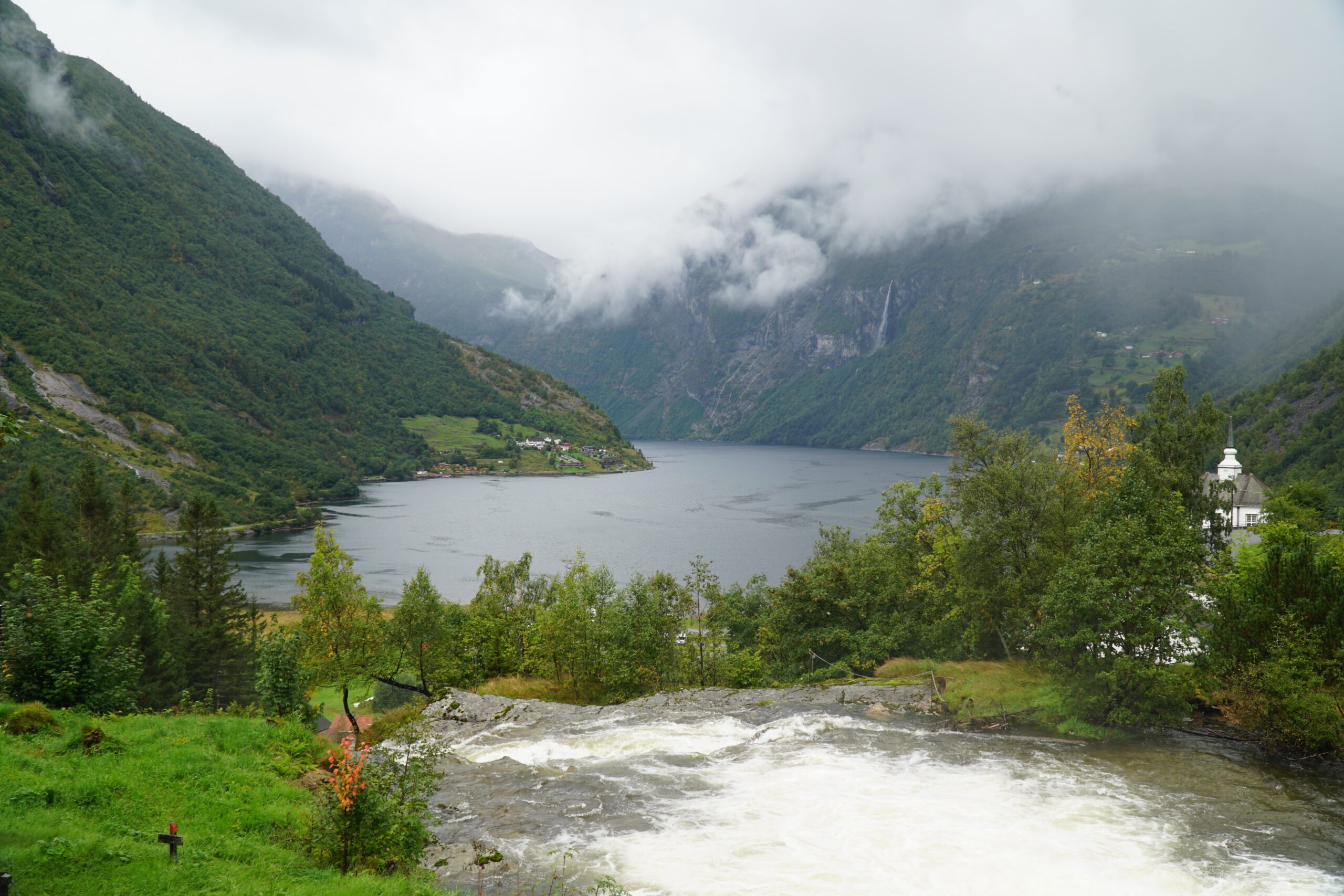 Panoramic view from Norsk Fjordsenter Geiranger overlooking the UNESCO World Heritage Geirangerfjord with cascading waterfall in foreground, green valley and misty Norwegian mountains