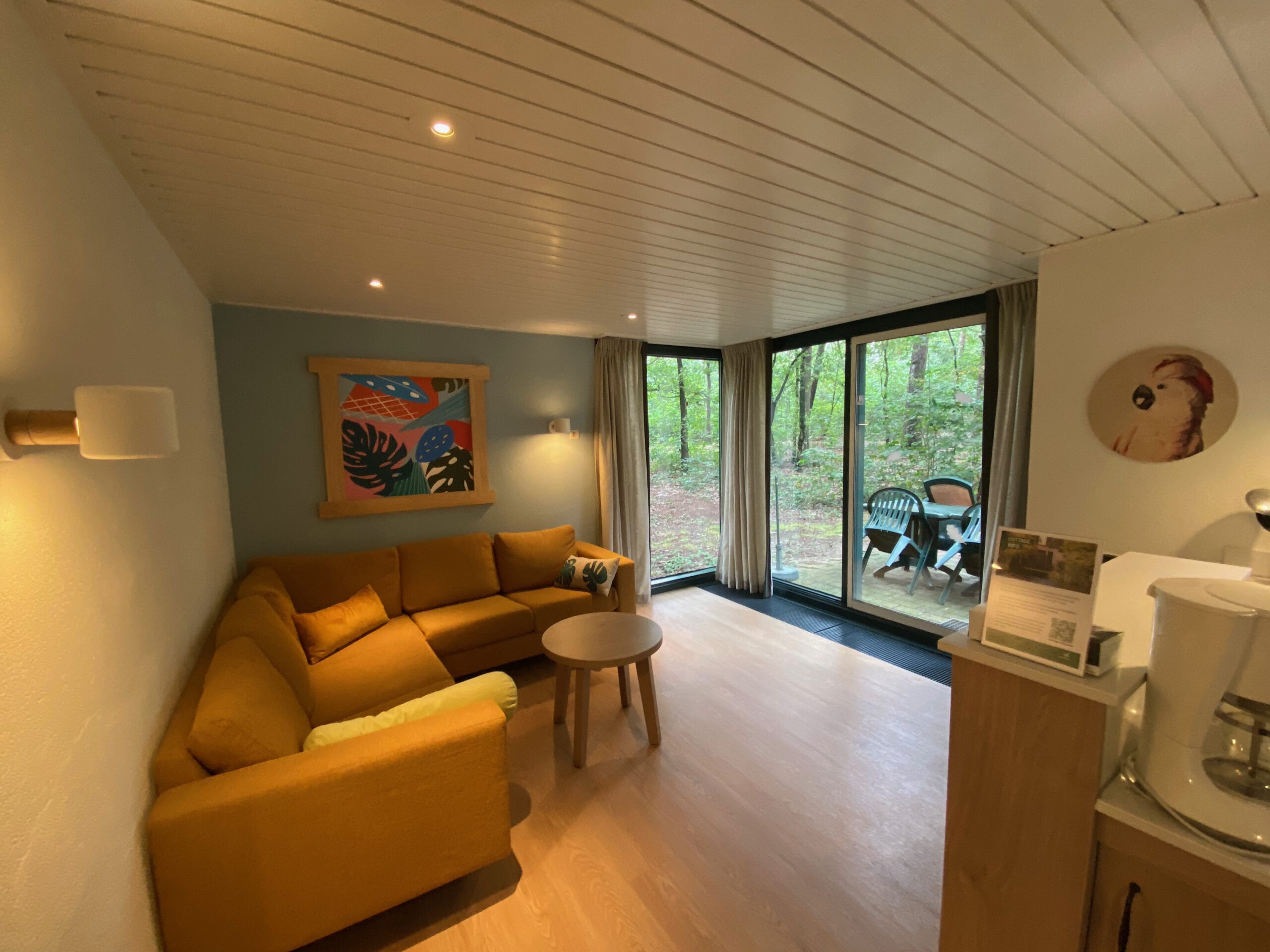 Living room inside Center Parcs Het Heijderbos 4 pers. Premium HB1922 cottage 965 featuring modern corner sofa, small coffee table and large windows with forest edge terrace view.