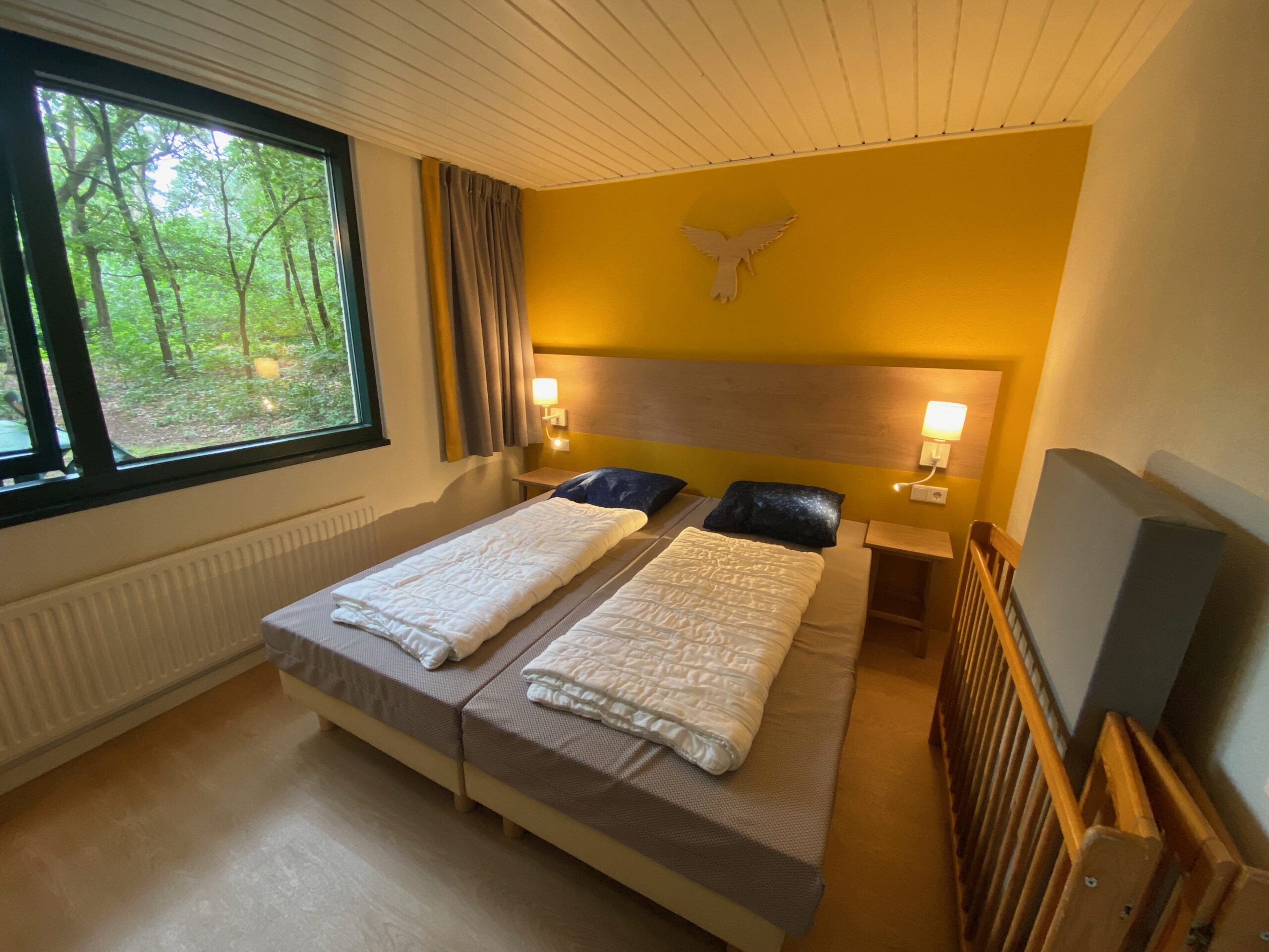 Bedroom inside Center Parcs Het Heijderbos 4 pers. Premium HB1922 cottage 965 featuring twin beds, yellow accent wall, bedside lamps and forest edge window view.