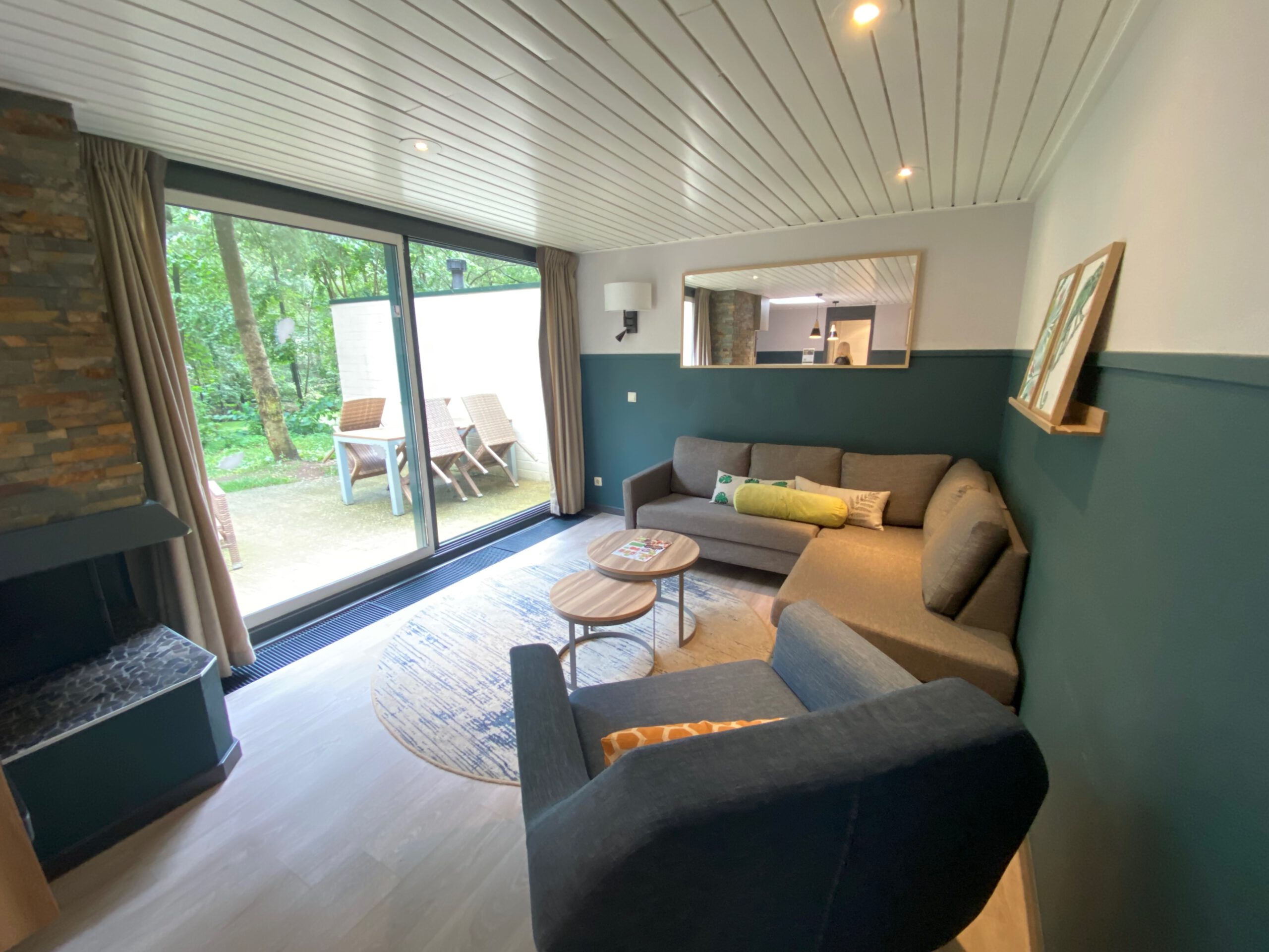 Spacious lounge area inside Center Parcs Het Heijderbos VIP cottage 978, featuring corner sofa, coffee tables, open fireplace, large panoramic windows and forest view terrace.