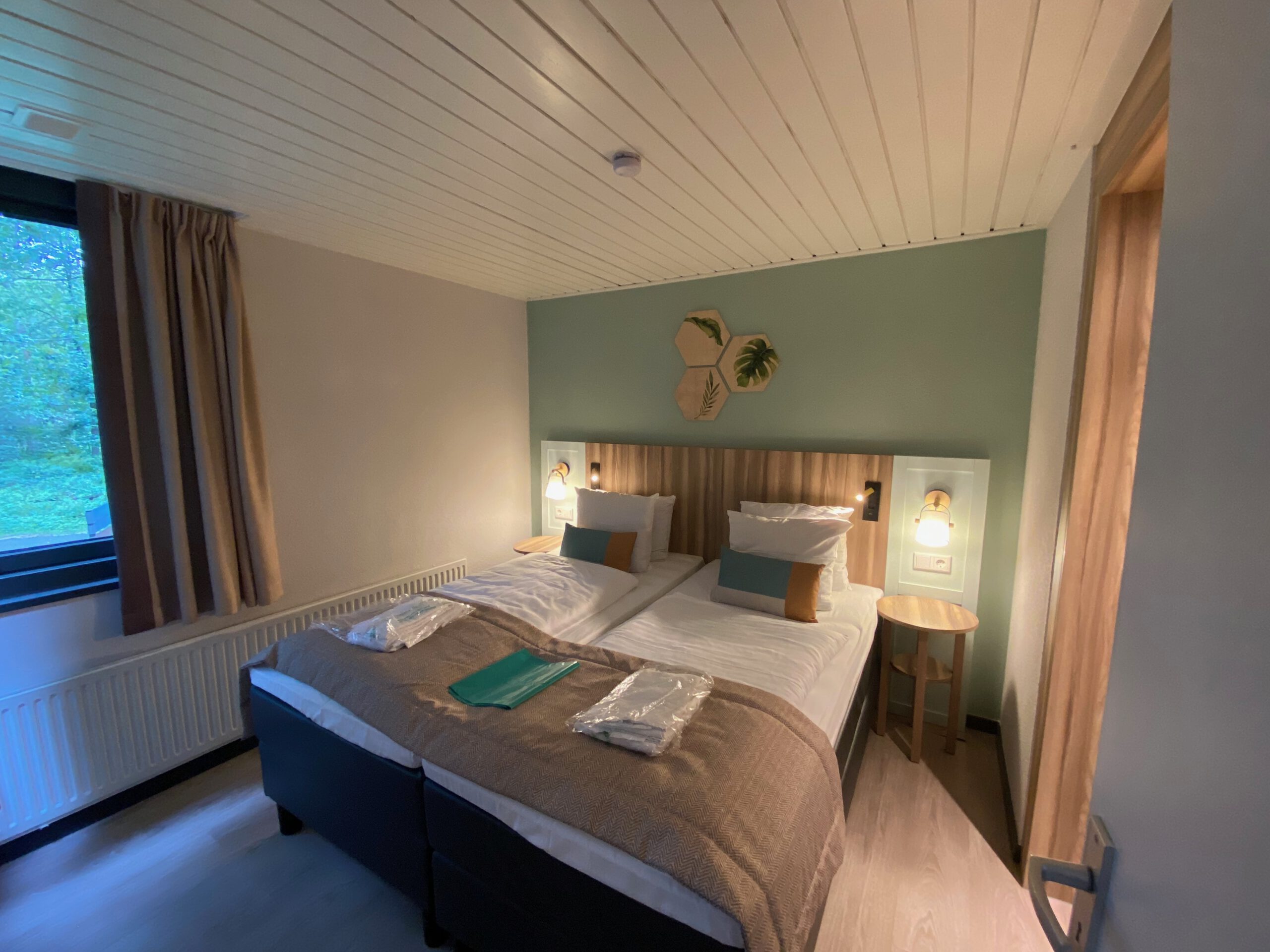 Second bedroom inside Center Parcs Het Heijderbos VIP cottage 978, featuring twin boxspring beds, bedside tables with reading lights, large radiator under the window and forest facing view.