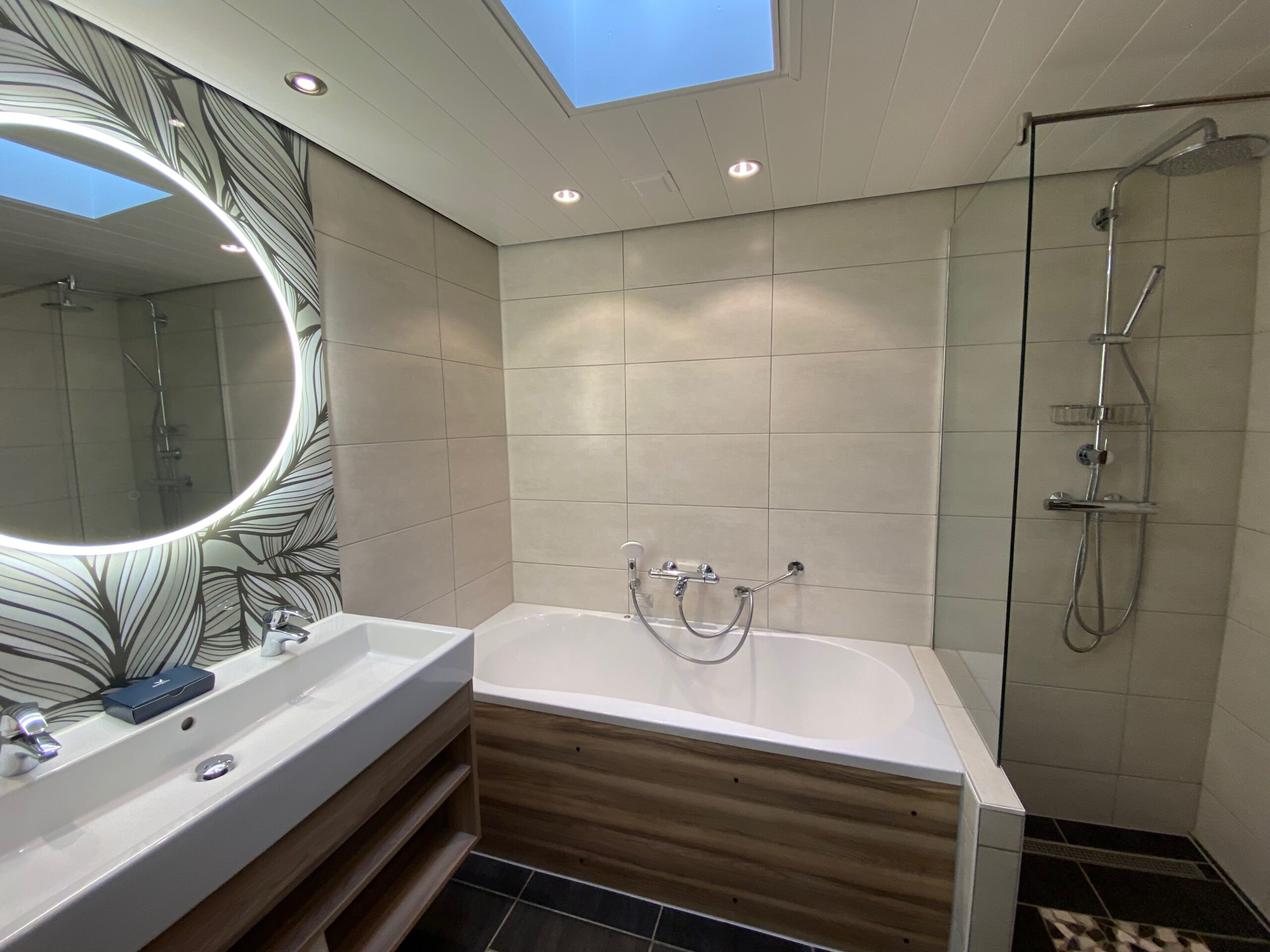 Luxury bathroom inside Center Parcs Het Heijderbos VIP cottage 978, featuring whirlpool bathtub, rain shower with glass wall, double sink with illuminated round mirror, skylight and high quality tile finish.