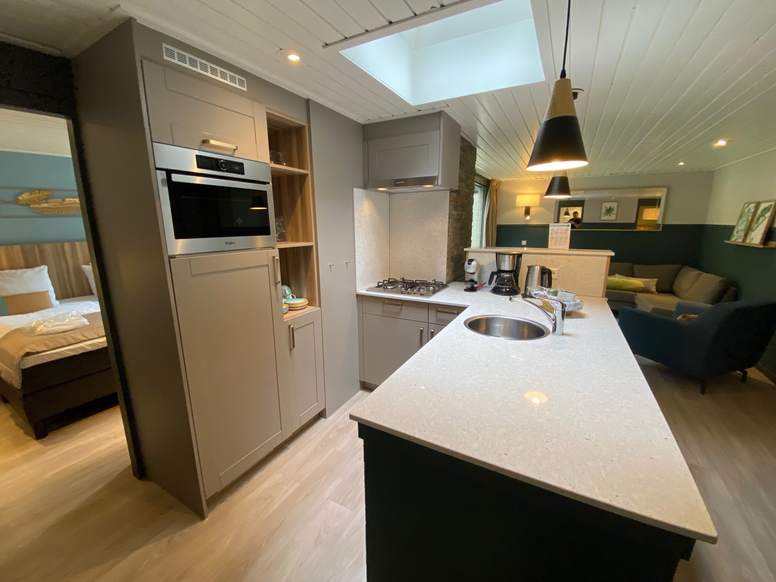 Luxury open kitchen and living area inside Center Parcs Het Heijderbos VIP cottage 978, featuring gas cooktop, built in combi microwave, natural stone wall finish, skylight, sink, Dolce Gusto machine, filter coffee maker, and direct view into the living room and bedroom.