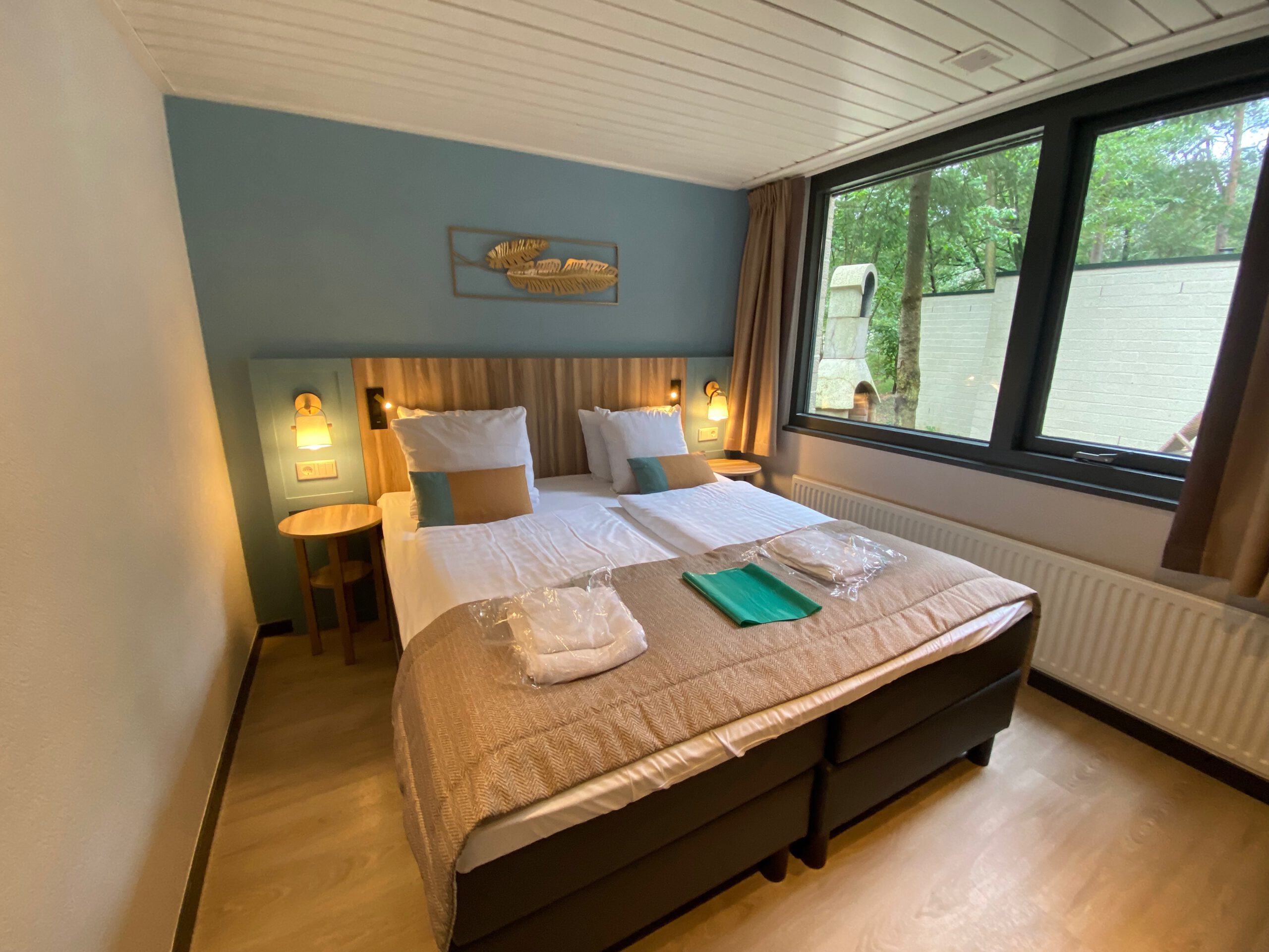 Master bedroom inside Center Parcs Het Heijderbos VIP cottage 978, featuring twin boxspring beds, bedside tables with dual wall lights, large window with forest view and direct sight on the private terrace.