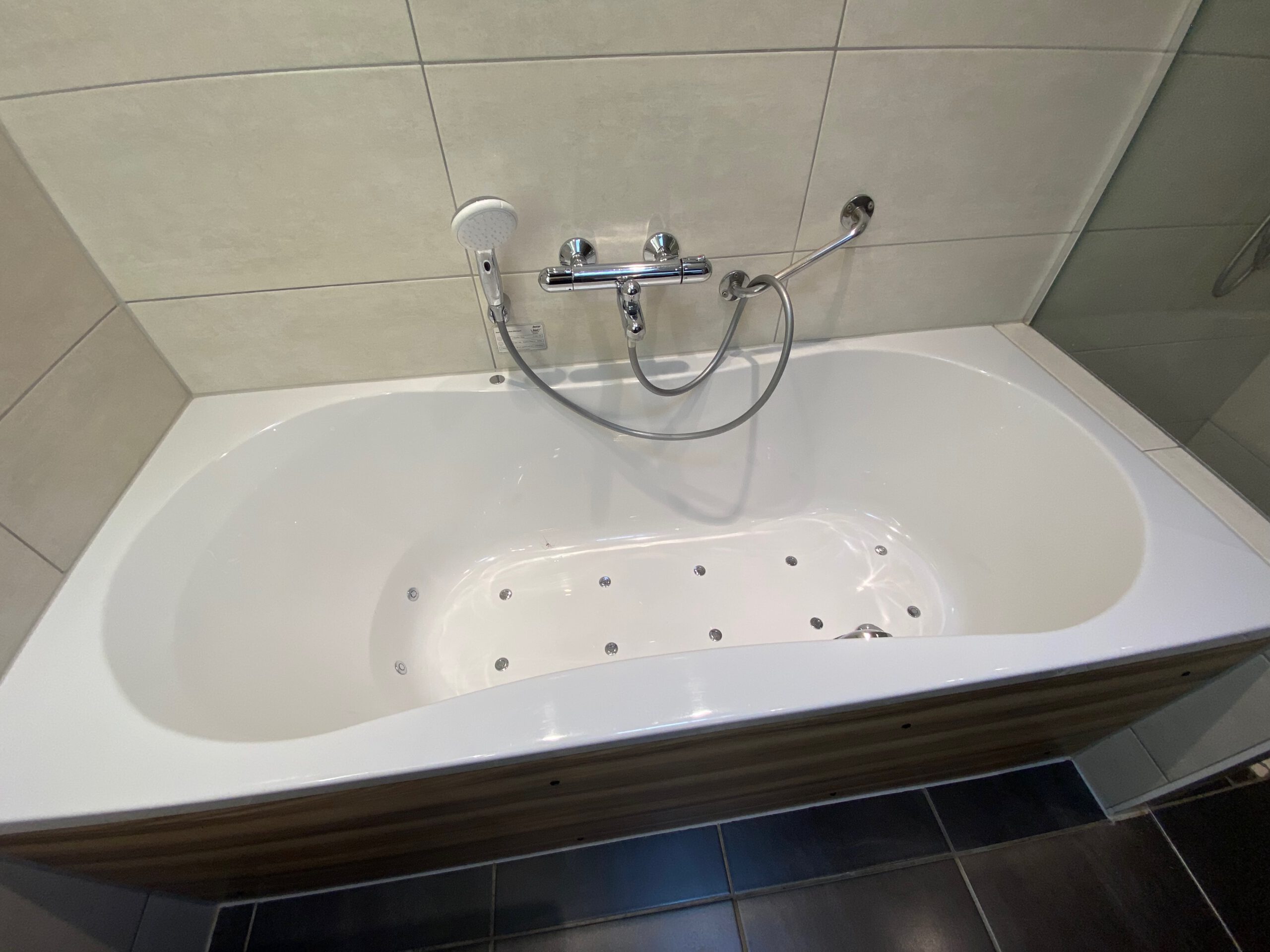 Whirlpool bathtub inside Center Parcs Het Heijderbos VIP cottage 978, featuring built in massage jets, handheld shower head, tiled walls and wooden panel finish.
