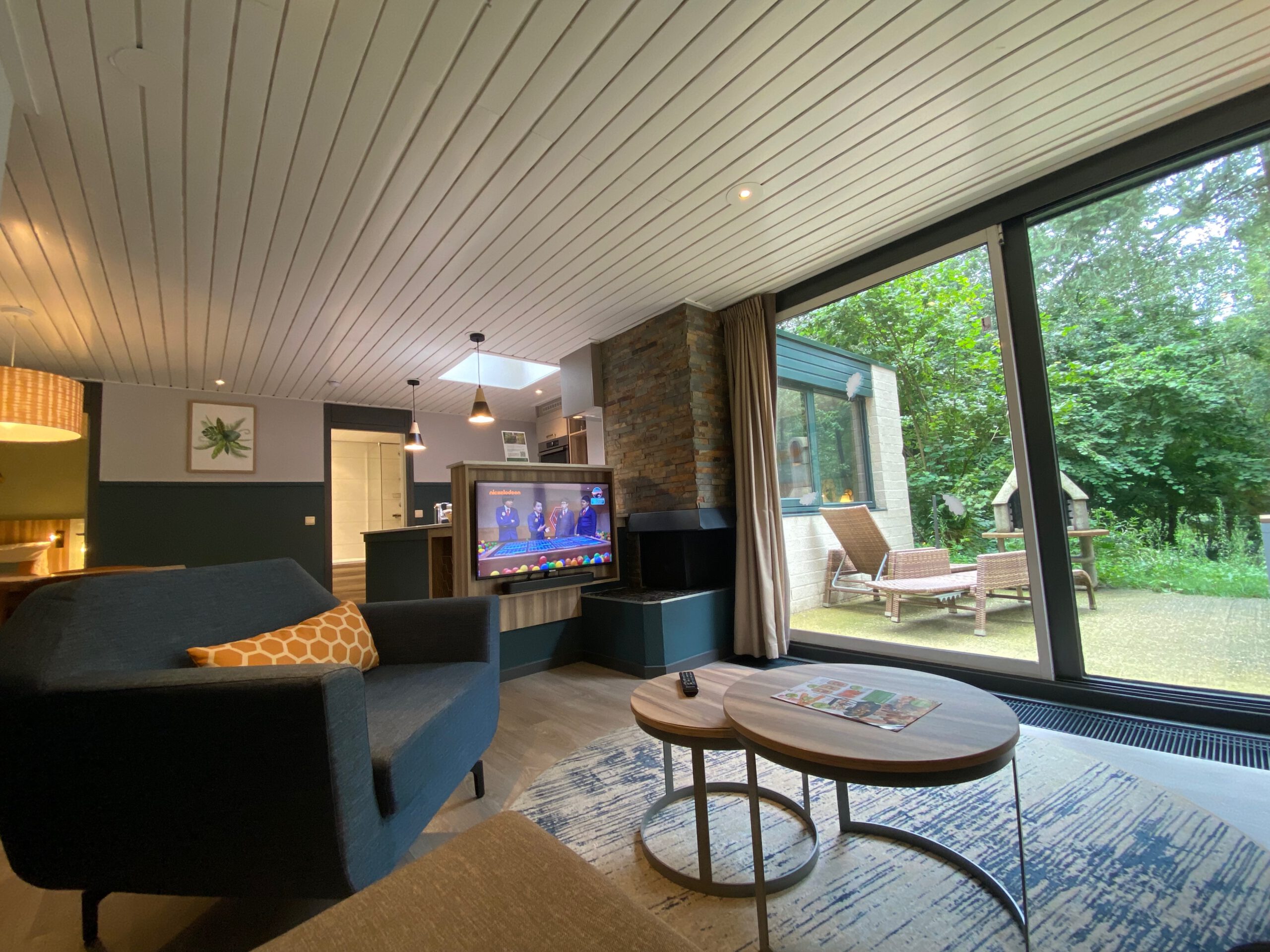 Modern living room inside Center Parcs Het Heijderbos VIP cottage 978, featuring comfortable sofa, armchair, open fireplace with natural stone wall, television, large sliding glass doors and direct access to the private forest terrace.
