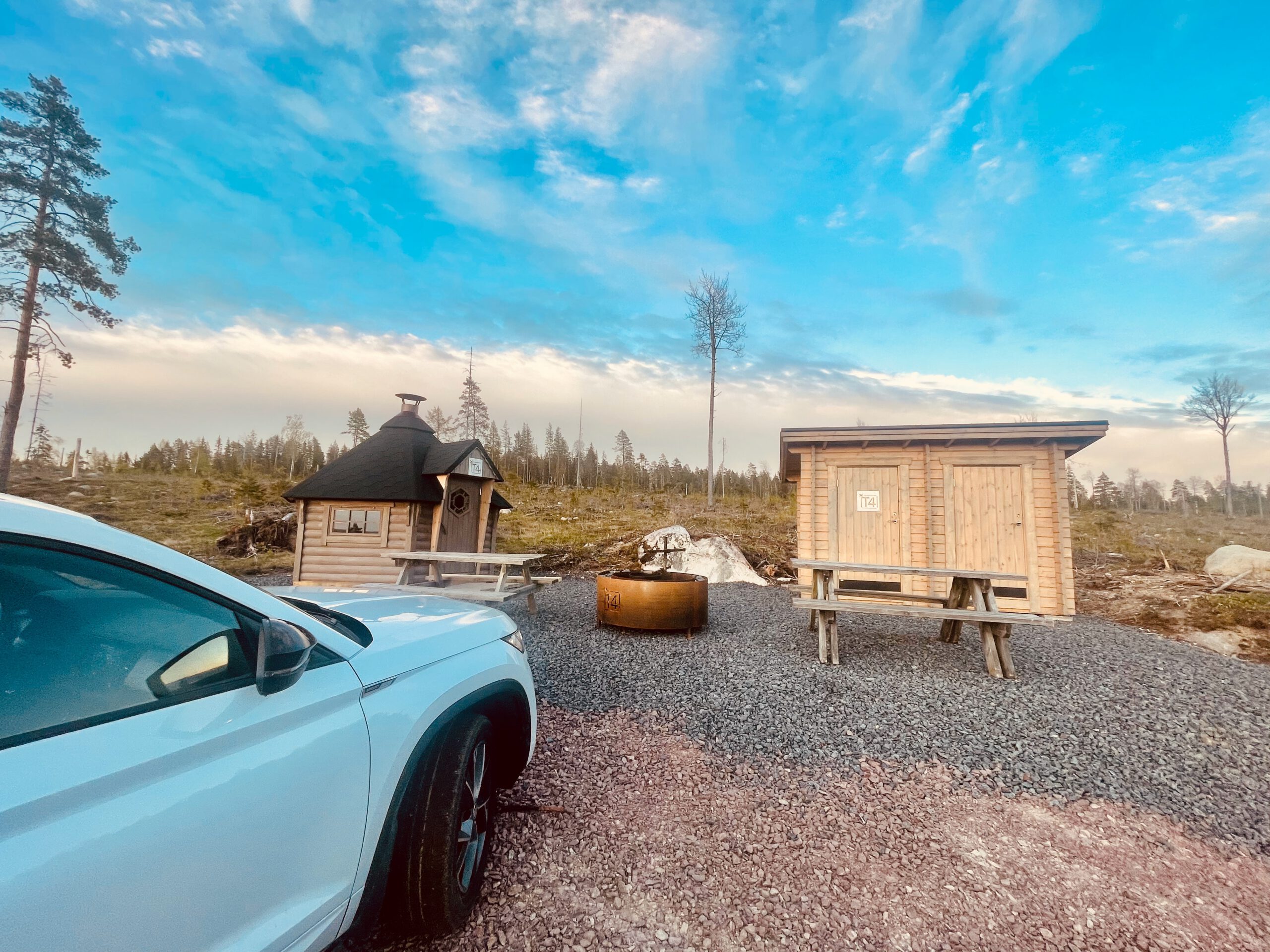 T4 Wintertrail Kota Sweden deep forest shelter area with traditional kota huts, fire pit and a white Skoda SUV parked off road in northern Sweden wilderness