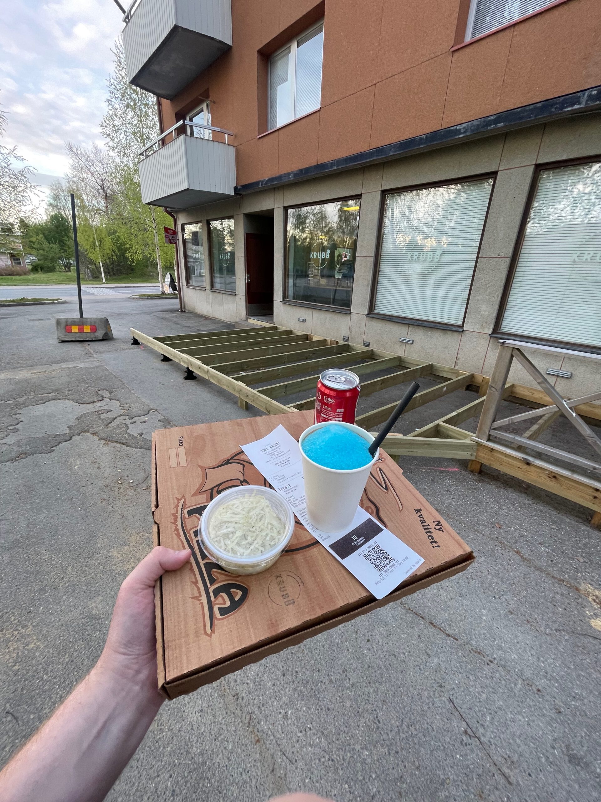 Takeaway order from Töre KRUBB restaurant in Töre Sweden, showing a pizza box, fresh slaw, receipt, Coca Cola and blue slush enjoyed outside the restaurant with the terrace under construction