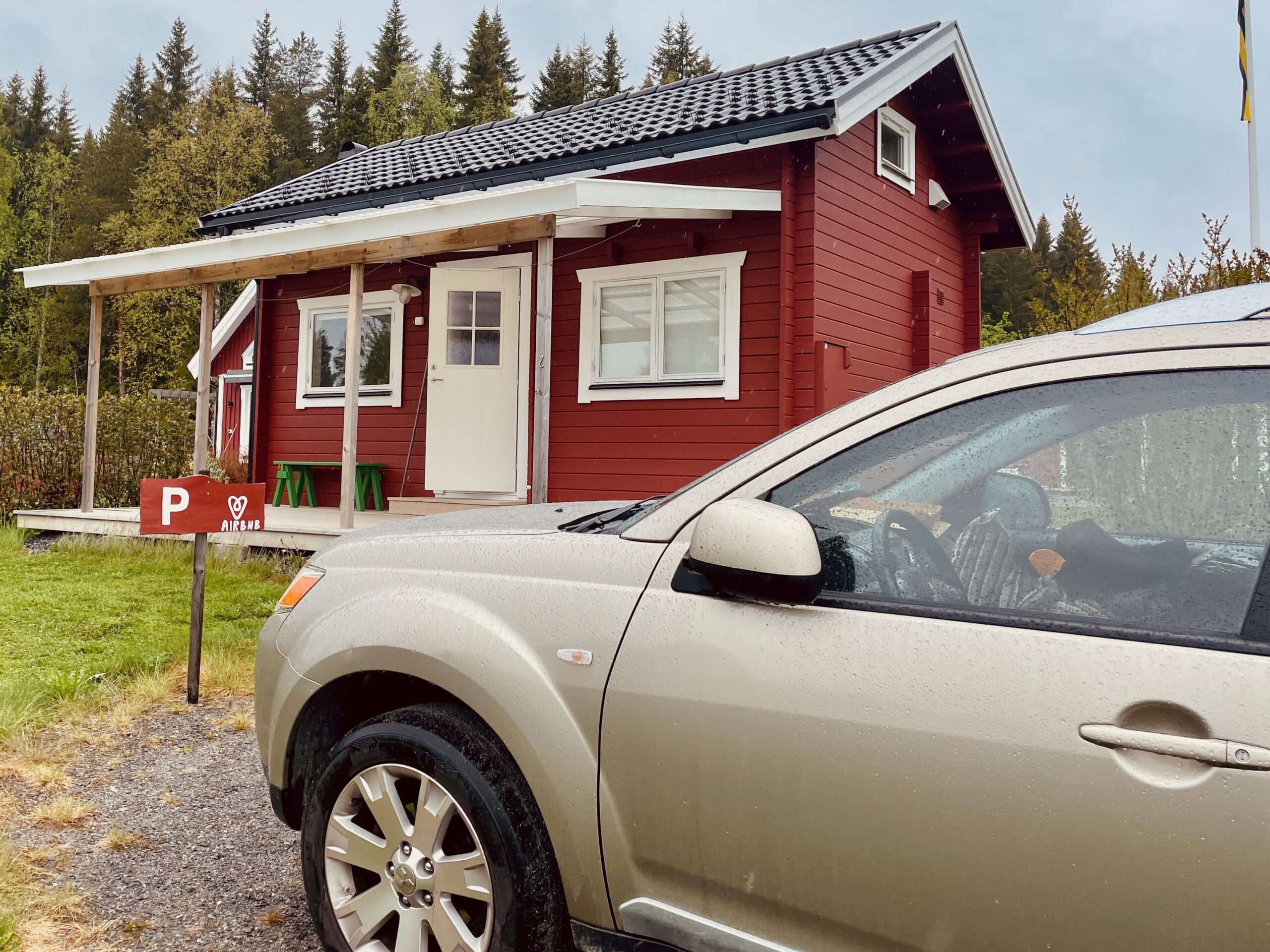 Cozy cottage with lake view Norra bergfors, red wooden Airbnb cottage in Sweden with private parking, forest surroundings and a car parked in front on a rainy day
