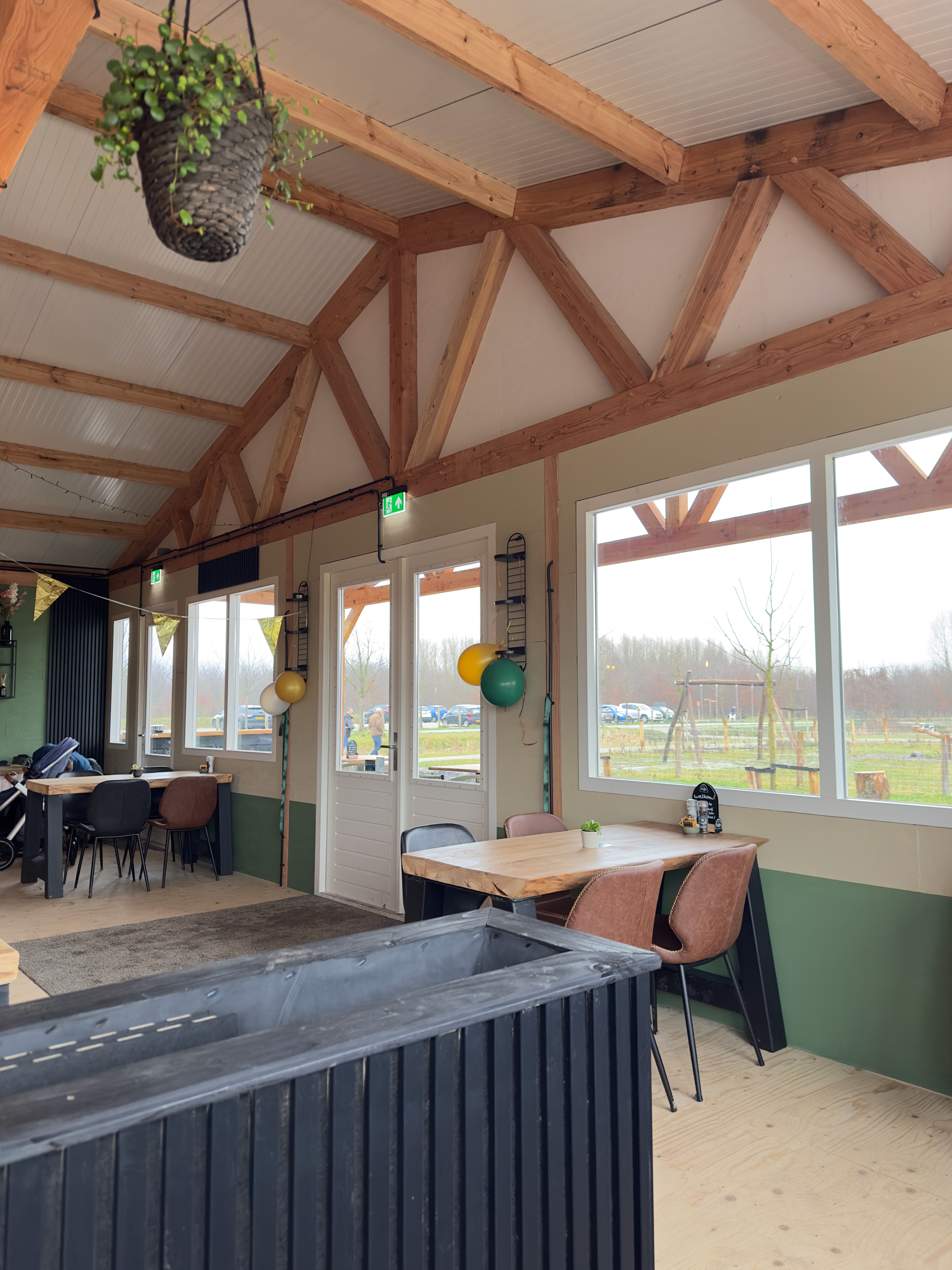 Bright seating area inside BentGoud Bentwoud with large windows, wooden tables, green accents, and views toward the Bentwoud nature area