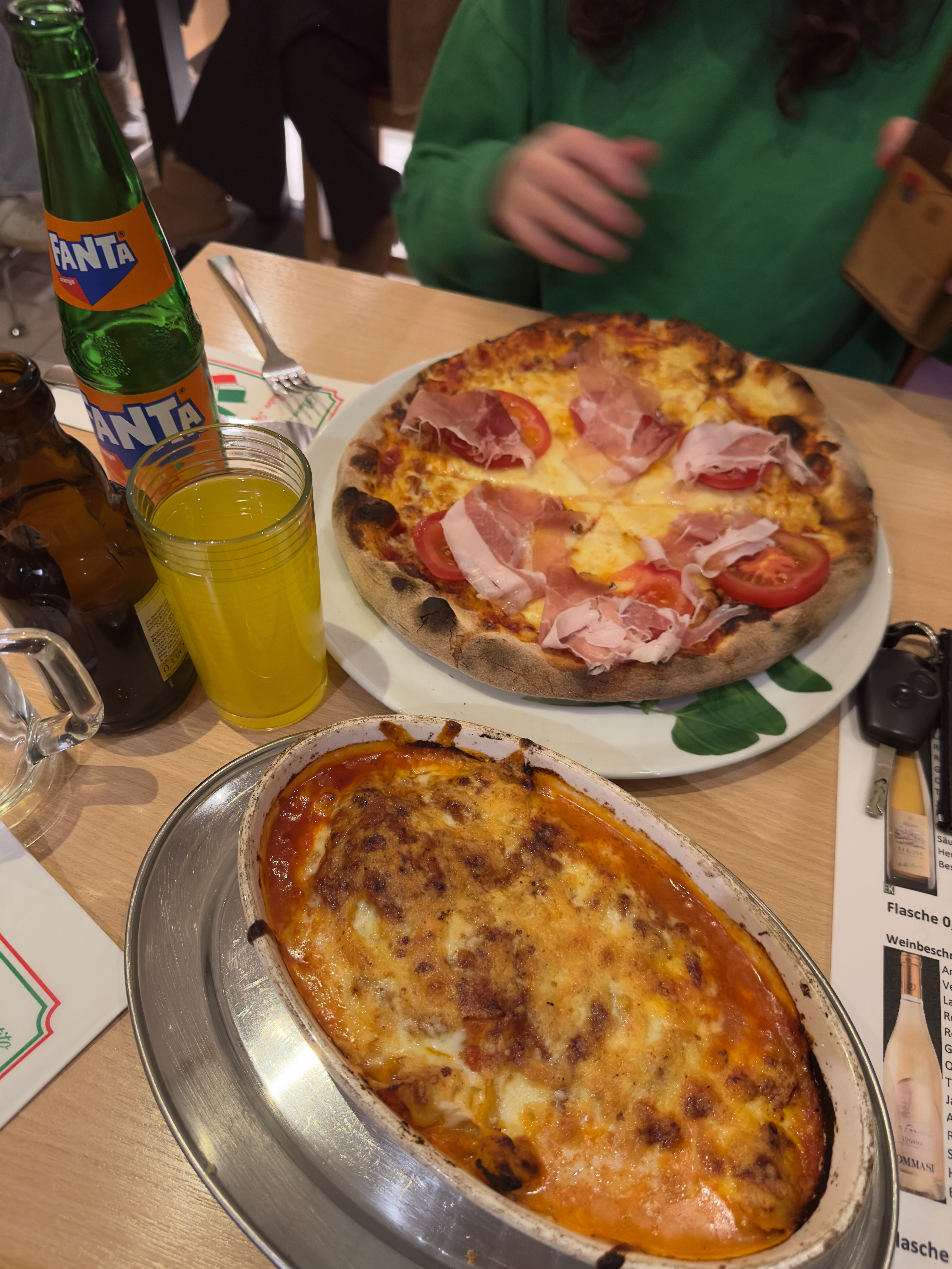 Pizzeria Il Genio Karken restaurant review showing a freshly served Italian pizza topped with prosciutto and tomatoes next to a baked lasagna dish, photographed during dinner inside the restaurant.