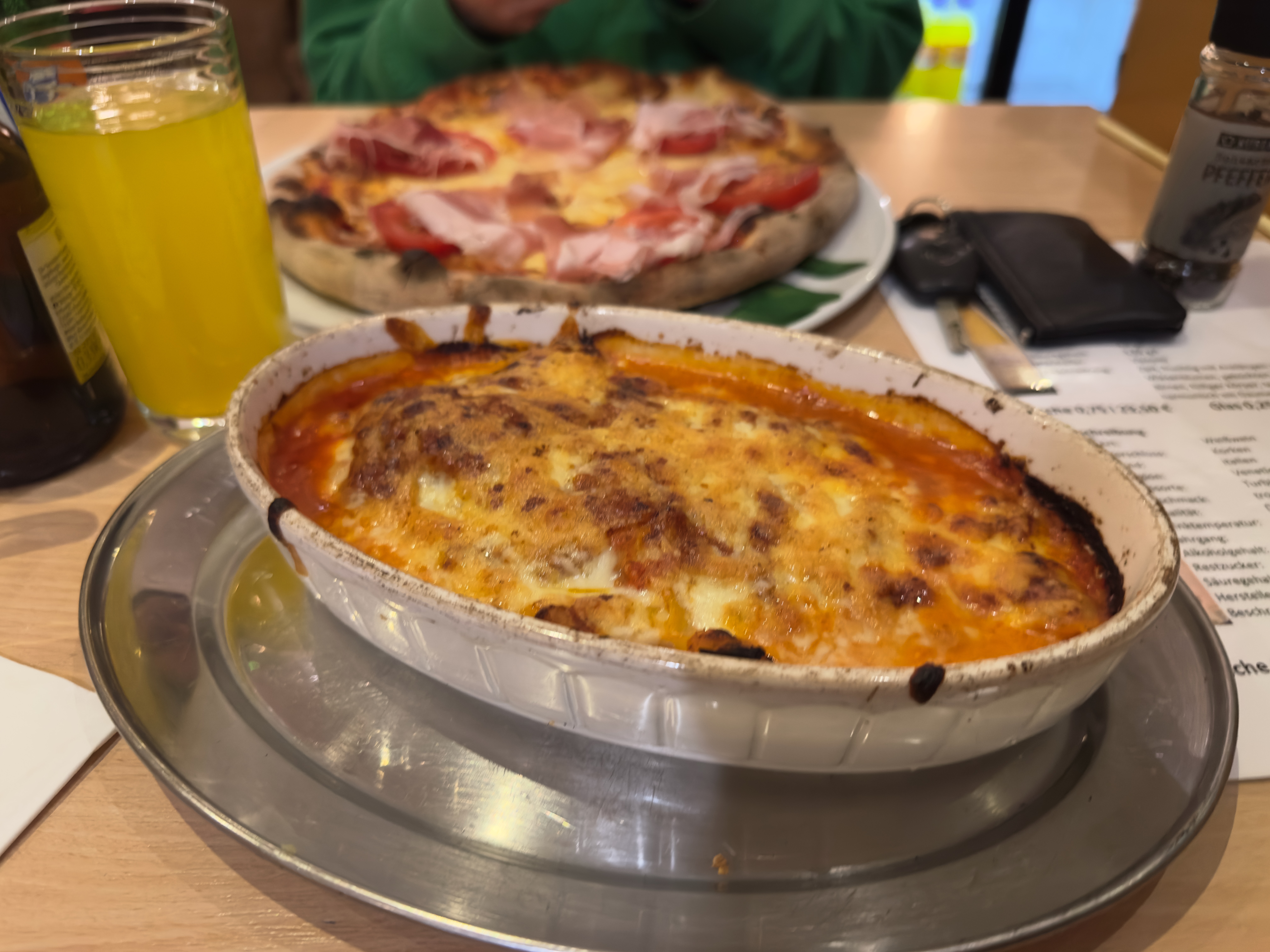 Pizzeria Il Genio Karken restaurant review showing a freshly baked Italian lasagna with tomato sauce, melted cheese, and a golden oven-baked crust, served hot on a metal plate with pizza and drinks visible in the background.