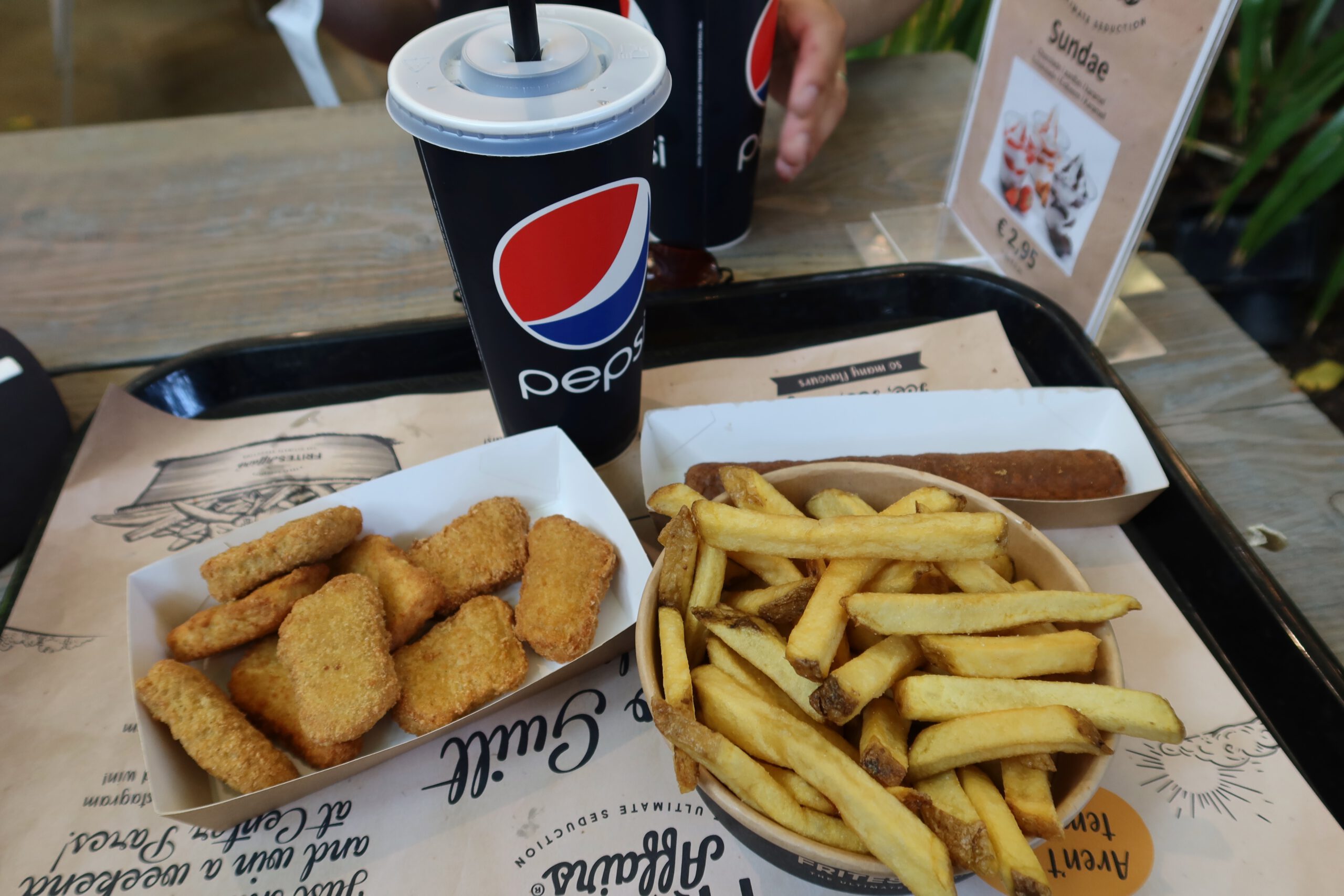 Center Parcs De Huttenheugte Day Visit lunch at Friet Affairs in the Market Dome, tray with golden French fries in a bowl, crispy breaded chicken nuggets, a fried snack roll, and two large Pepsi soft drinks on a wooden table inside the Action Factory area in Drenthe.