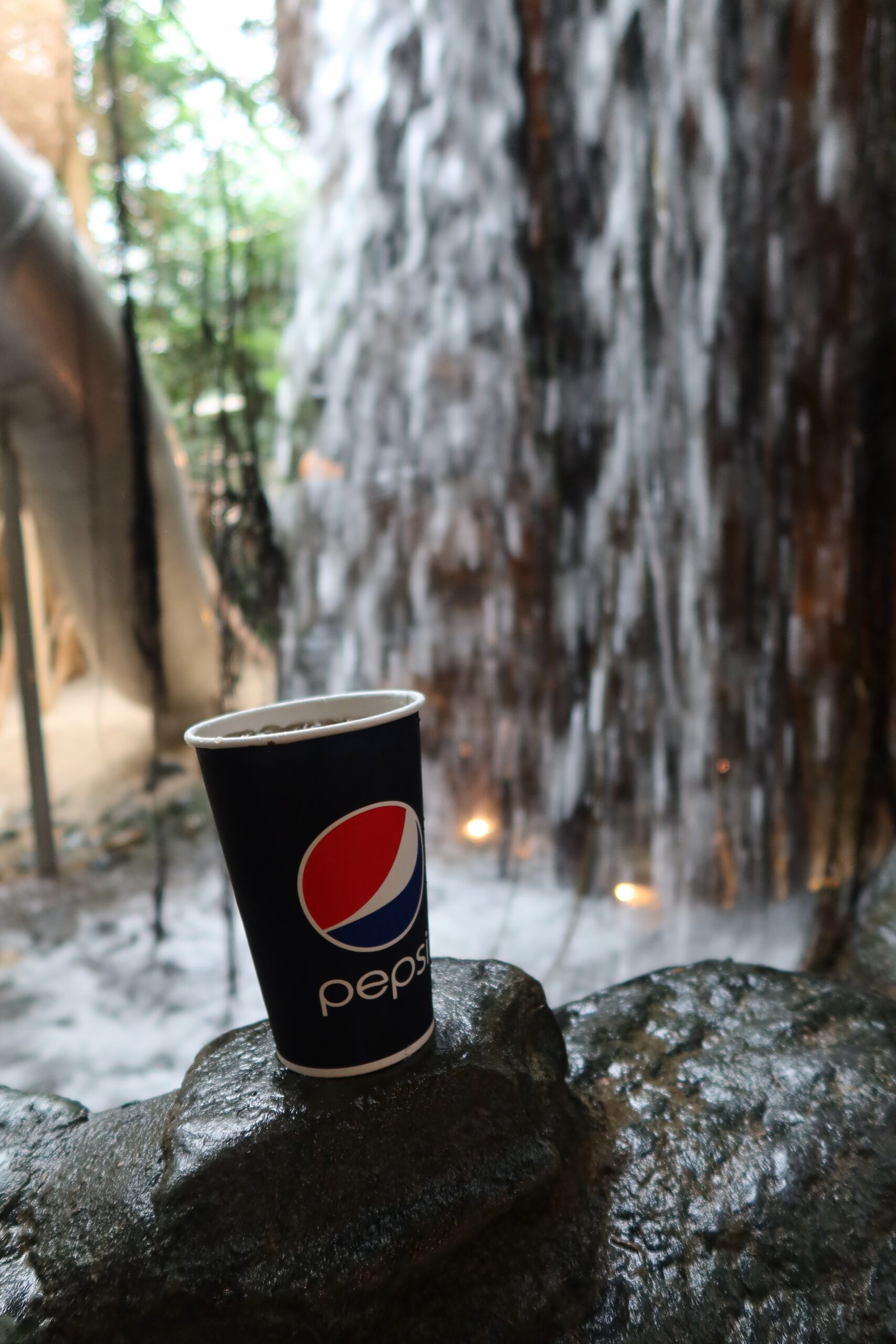 A cold Pepsi Cola drink in a paper cup at Center Parcs Het Heijderbos Jungle Dome, tropical indoor setting with waterfall and lush greenery in Limburg, The Netherlands.
