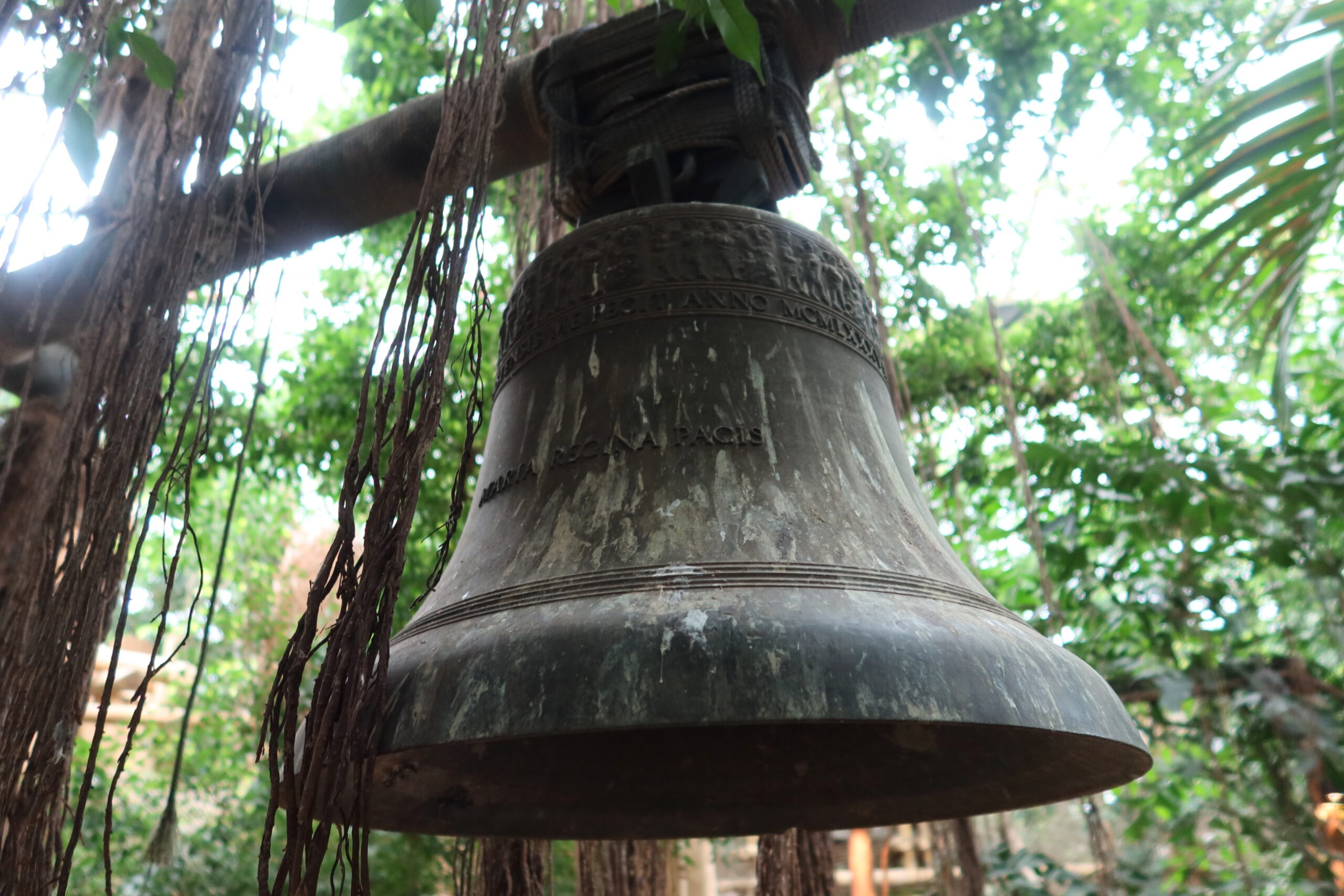 Large bronze jungle bell hanging among tropical trees inside the Jungle Dome Center Parcs Het Heijderbos, framed by dense indoor rainforest canopy.