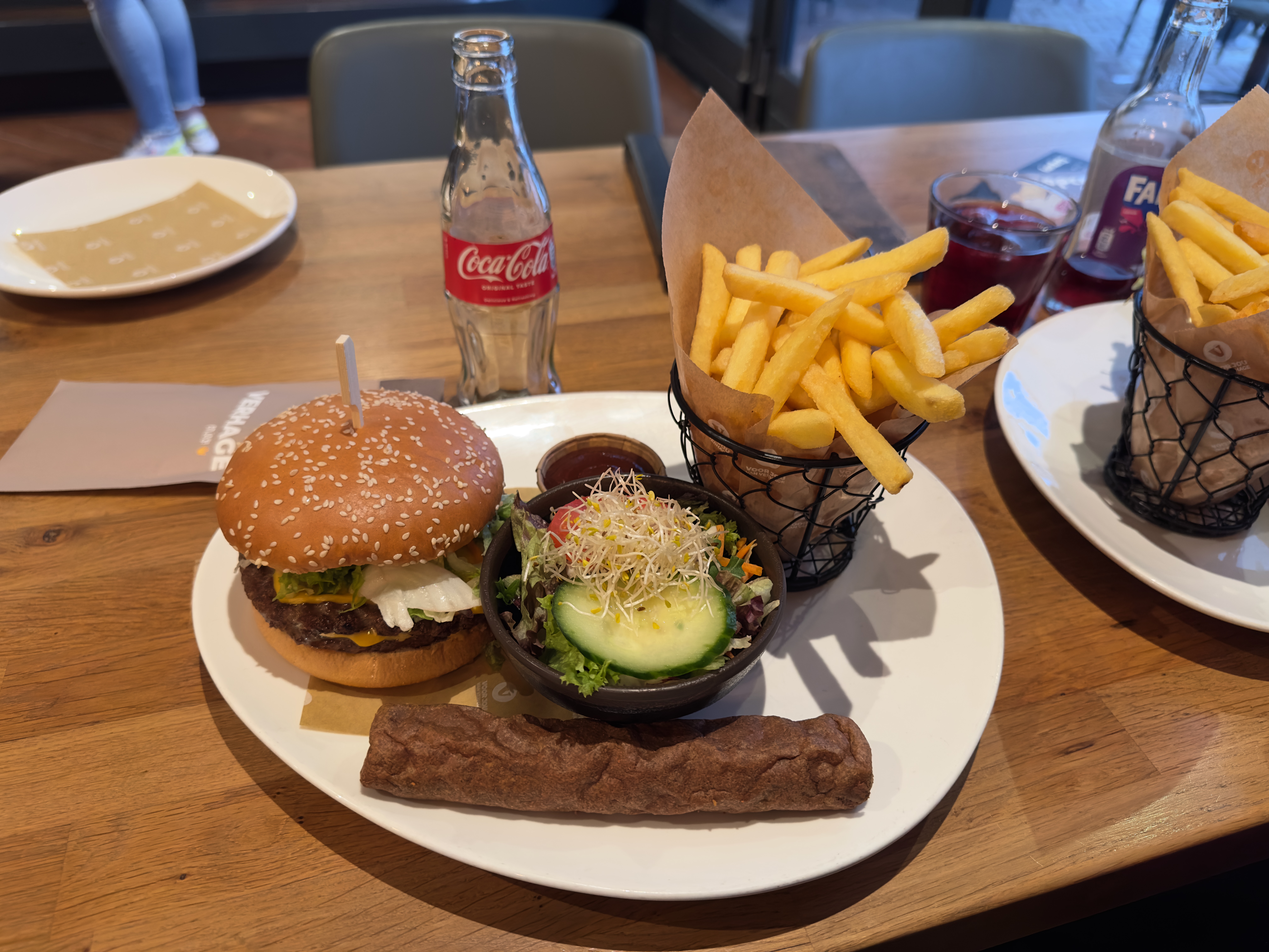 Verhage Burgh Haamstede double cheeseburger with sesame bun, fresh fries in basket, frikandel, side salad with cucumber and sprouts, and Coca Cola on wooden table inside the restaurant in Burgh Haamstede, Netherlands.