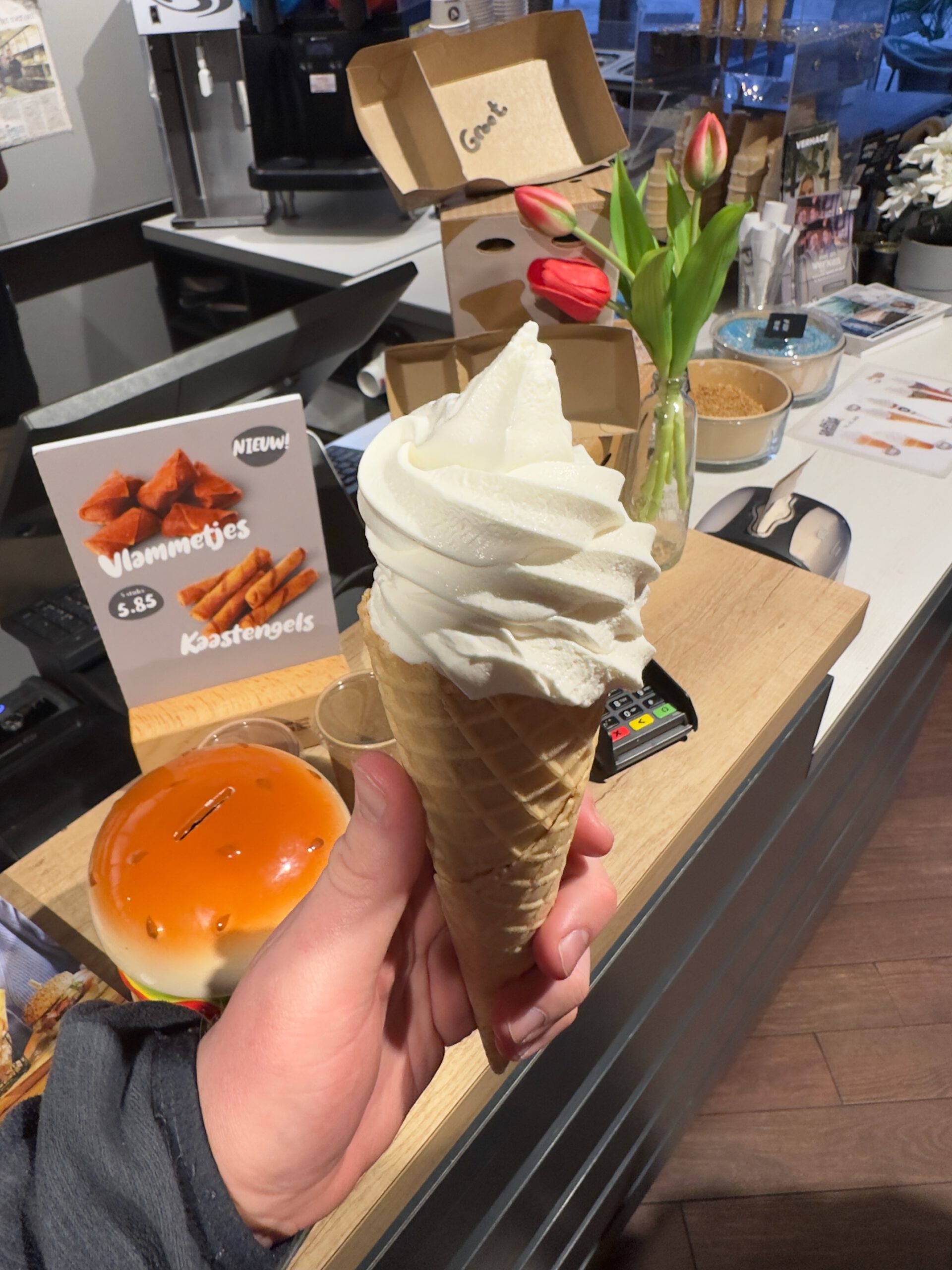 Verhage Burgh Haamstede soft serve ice cream in waffle cone held inside the restaurant, creamy vanilla swirl near the counter with snack display and menu sign in Burgh Haamstede, Netherlands.