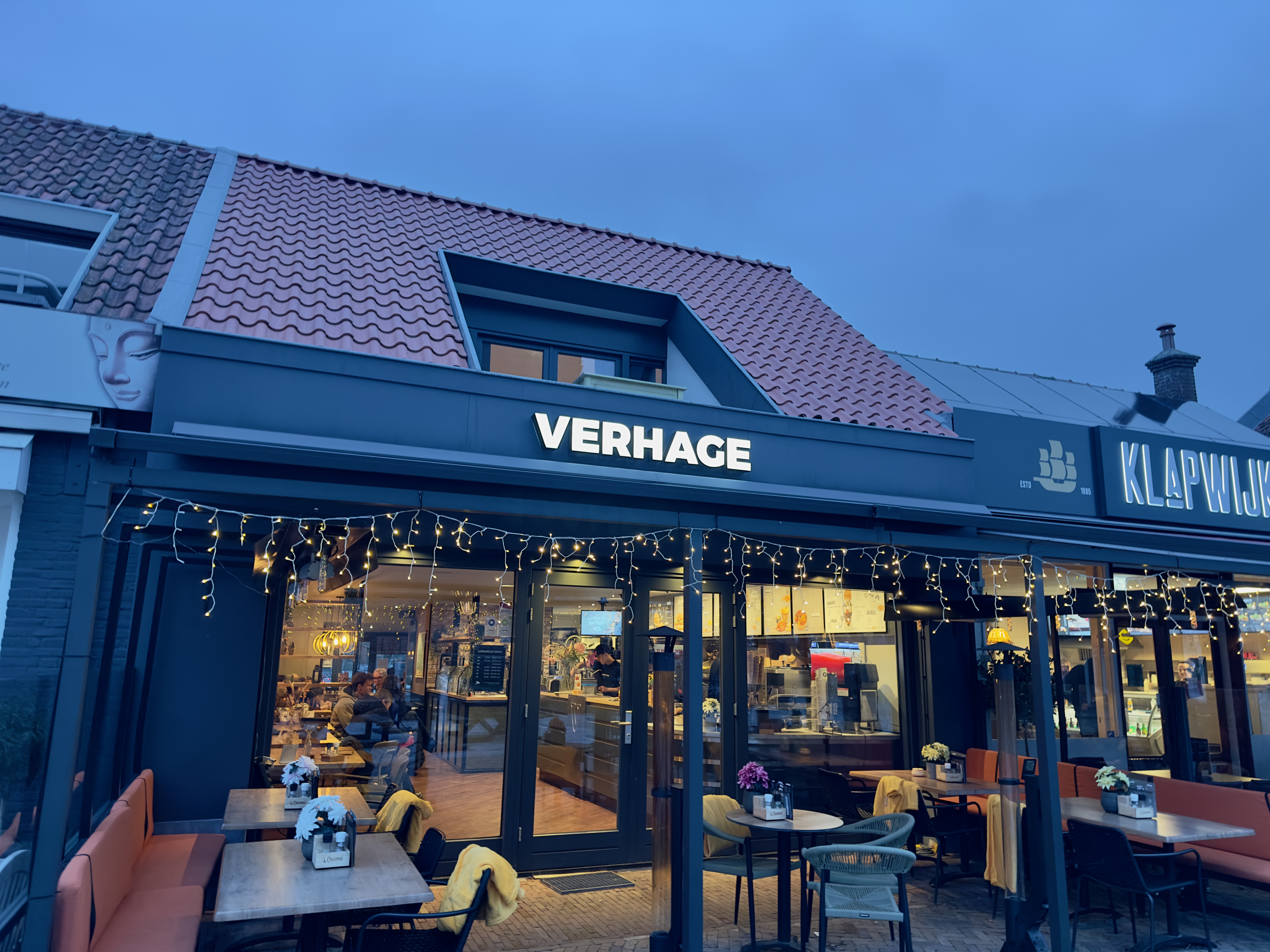 Verhage Burgh Haamstede restaurant exterior in the evening, modern dark facade with illuminated Verhage logo, cozy terrace seating with fairy lights, outdoor tables and chairs, and visible indoor dining area through large glass windows in Burgh Haamstede, Netherlands.