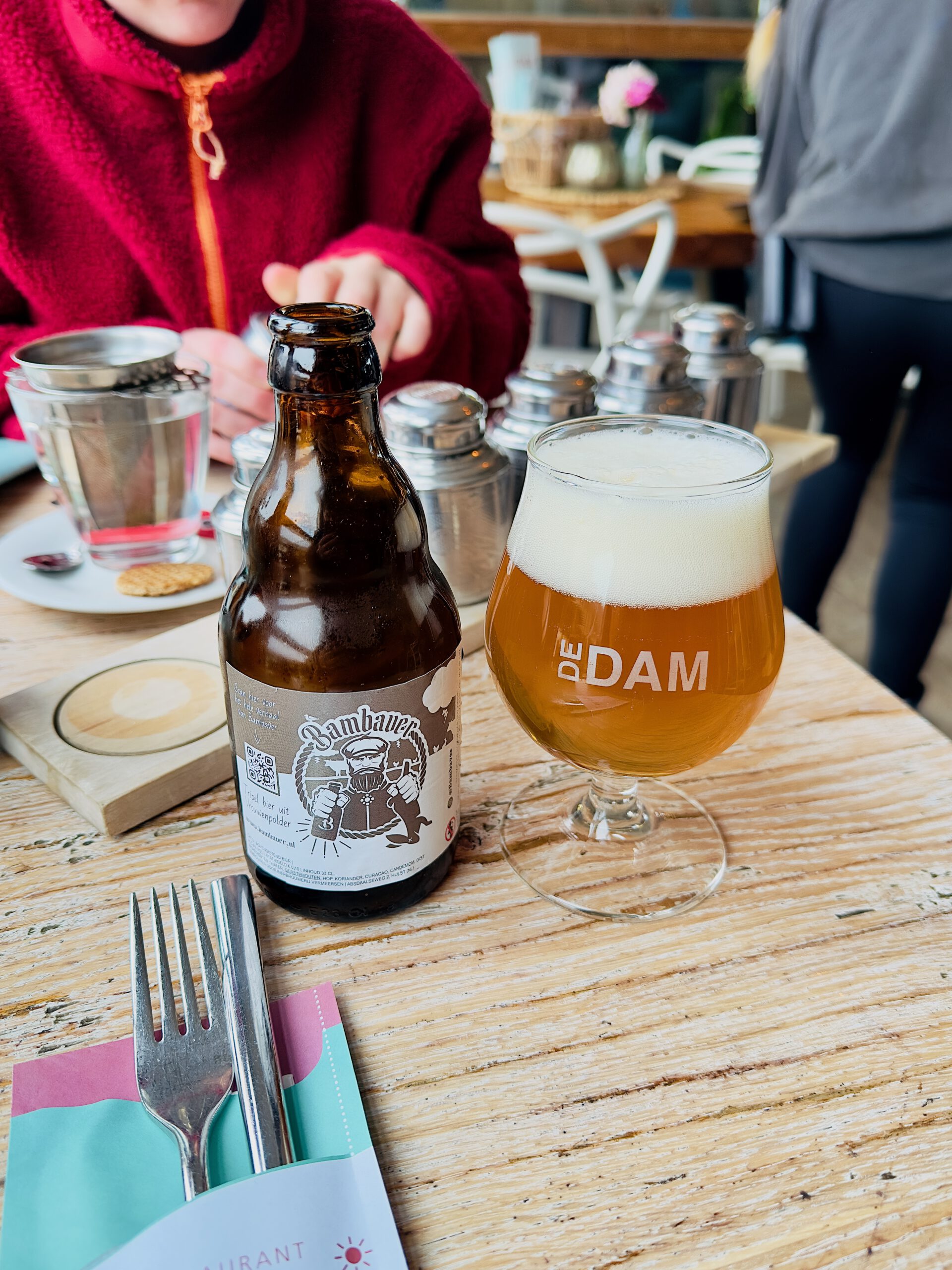 Strandrestaurant De Dam table setting with a Bambaver local beer bottle and a De Dam branded beer glass filled with golden beer and foam head, tea served in a glass cup with fresh herbs and strainer, cutlery in pastel colored napkin holder, wooden beach style table, person wearing red fleece jacket in the background and warm interior atmosphere inside the beach pavilion in Vrouwenpolder.