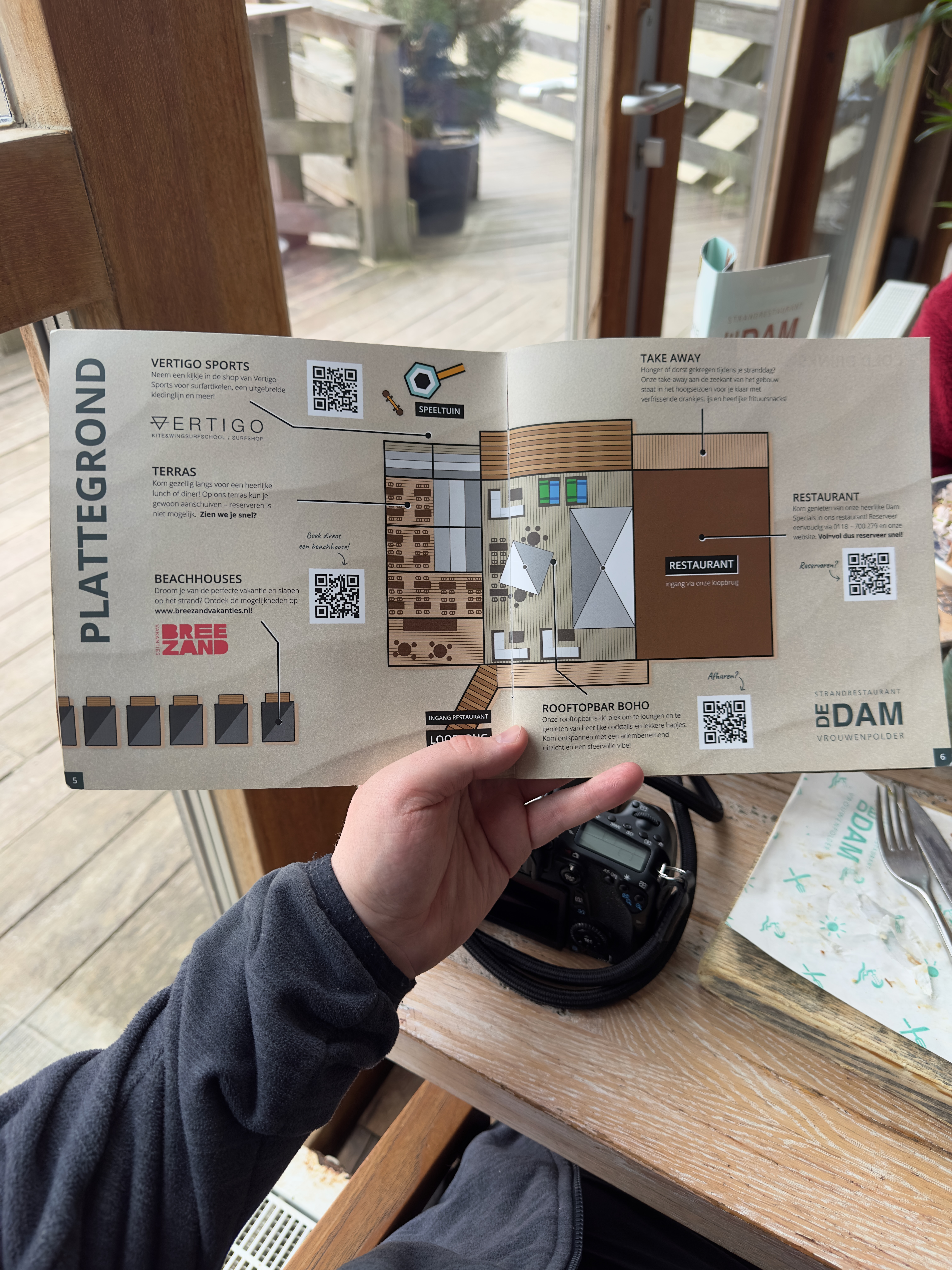 Strandrestaurant De Dam floor plan brochure held in hand inside the beach pavilion, showing illustrated layout with terrace, rooftop bar BOHO, take away area and restaurant entrance, QR codes printed on the map, wooden interior, glass door and deck outside visible in the background, camera body resting on the wooden table next to the brochure.