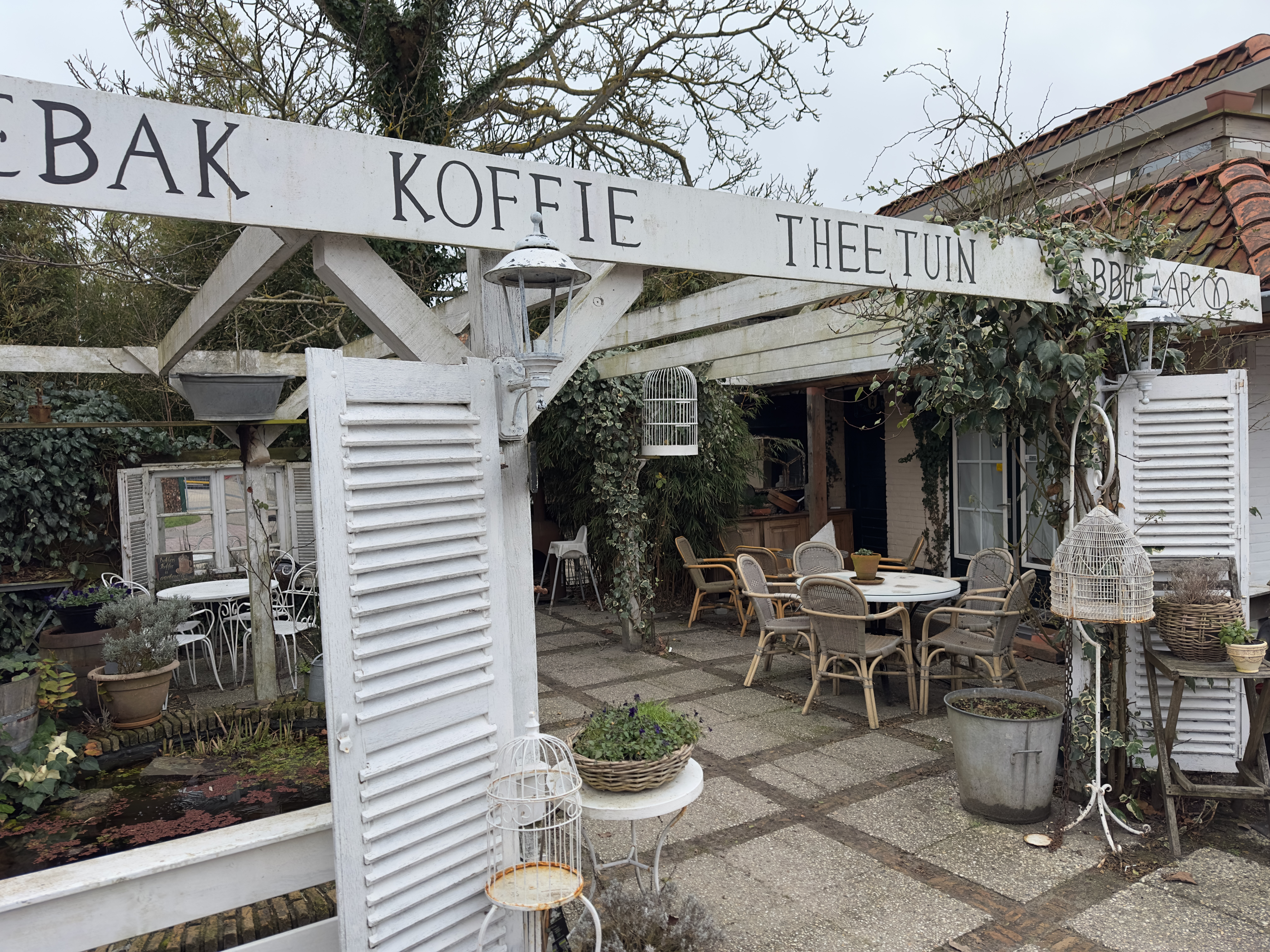 Bakkerij Sonnemans in Burgh Haamstede tea garden entrance with white wooden gate, coffee sign, ivy covered pergola and cozy outdoor seating area