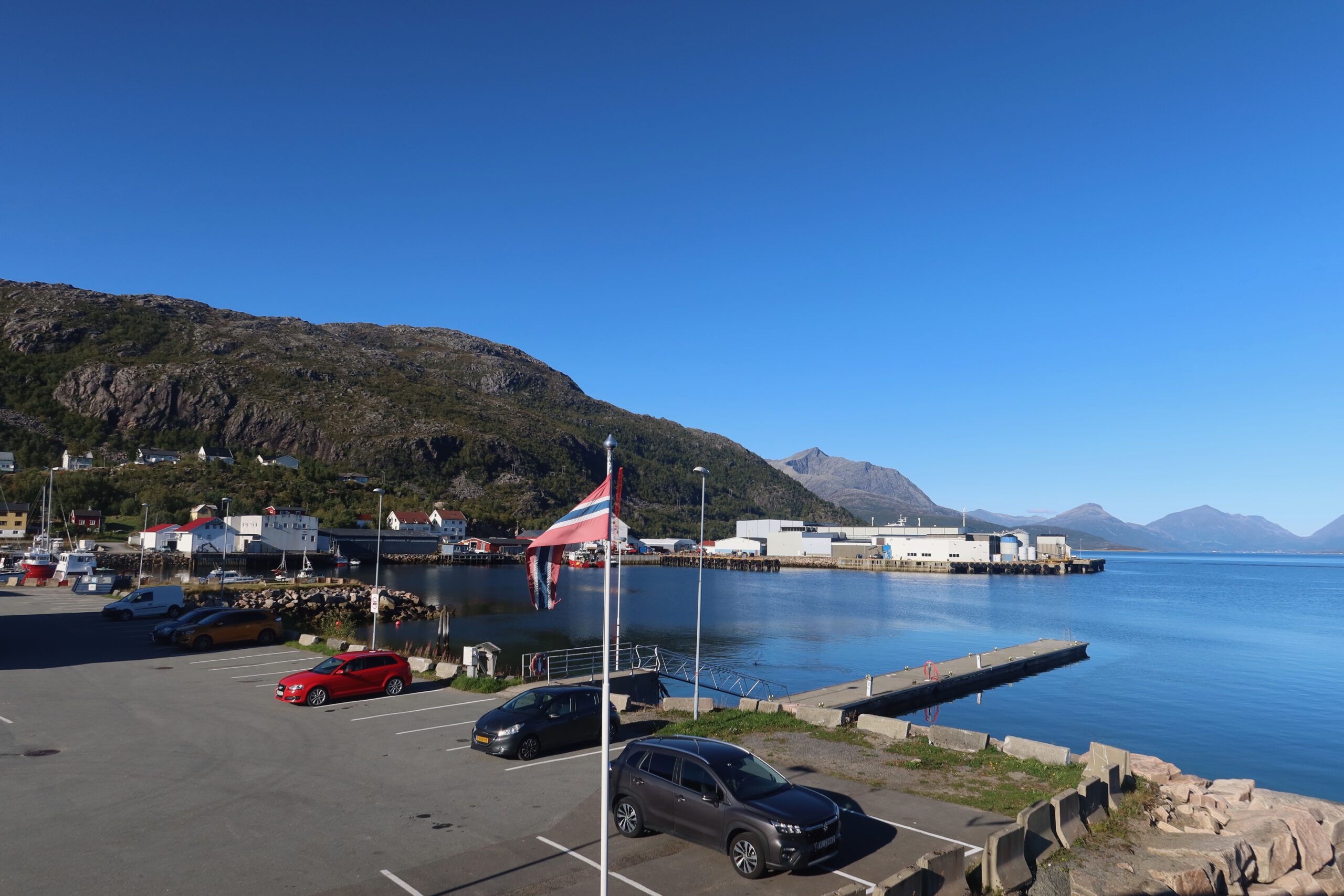 Lødingen Brygge Lofoten Norway parking area with Norwegian flag, harbor, calm fjord water and mountain scenery in the background