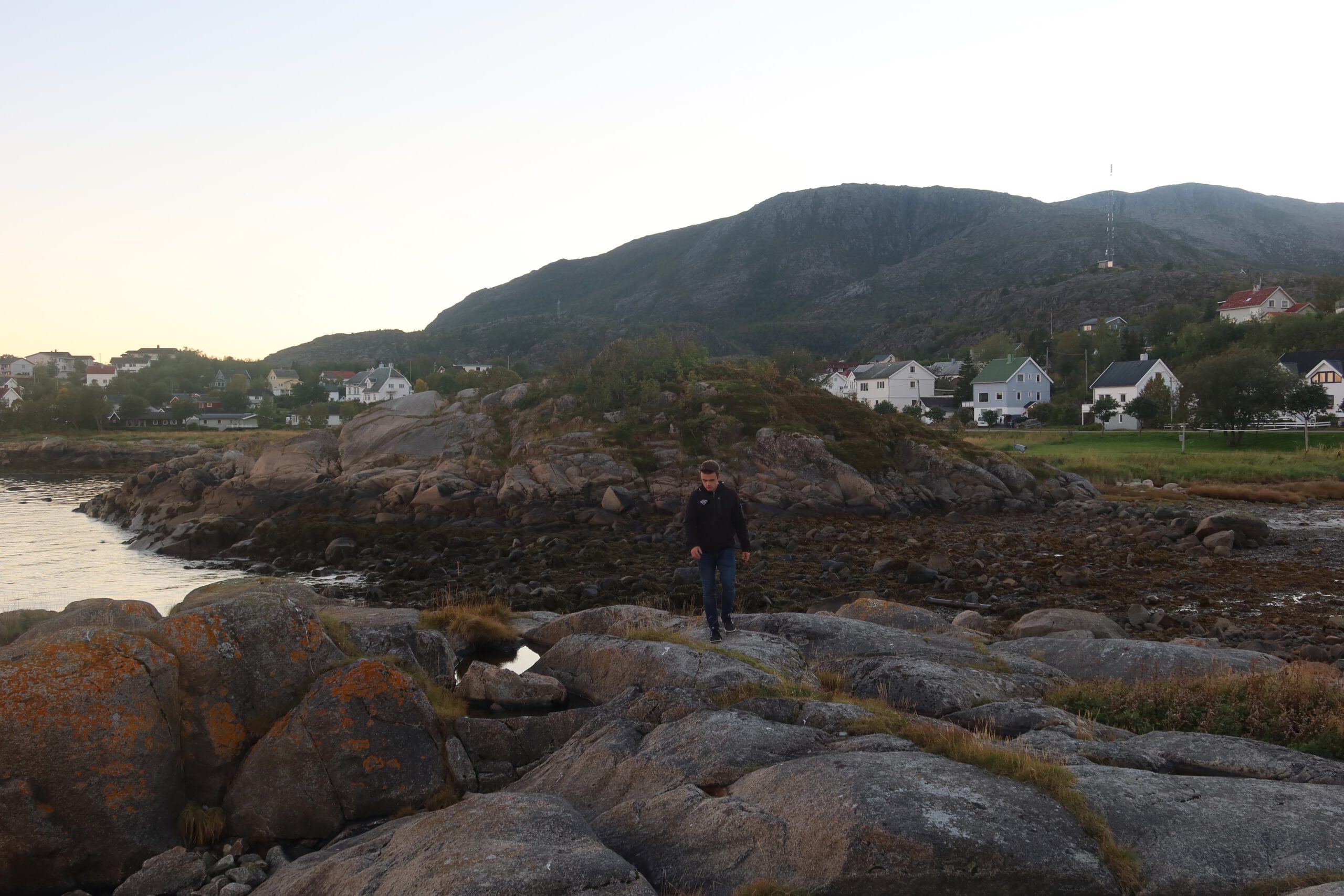Hjortholmen Northern Lights Spot during early evening light, featuring the rocky shoreline of Lødingen with coastal houses and mountains before nightfall, and Peter Smit walking