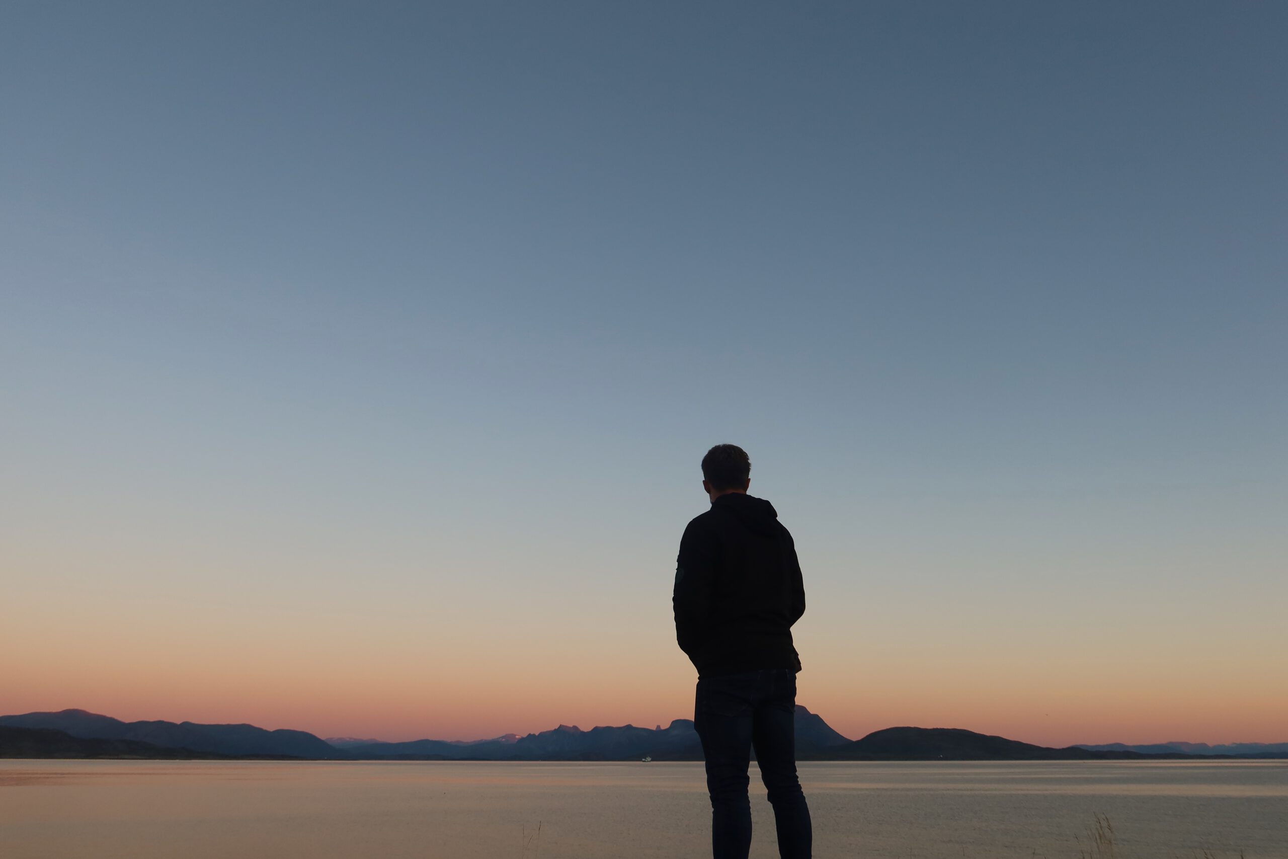 Hjortholmen Northern Lights Spot at dusk in Lødingen, showing Peter Smit standing by the calm sea with pastel Arctic sky colors and mountains on the horizon