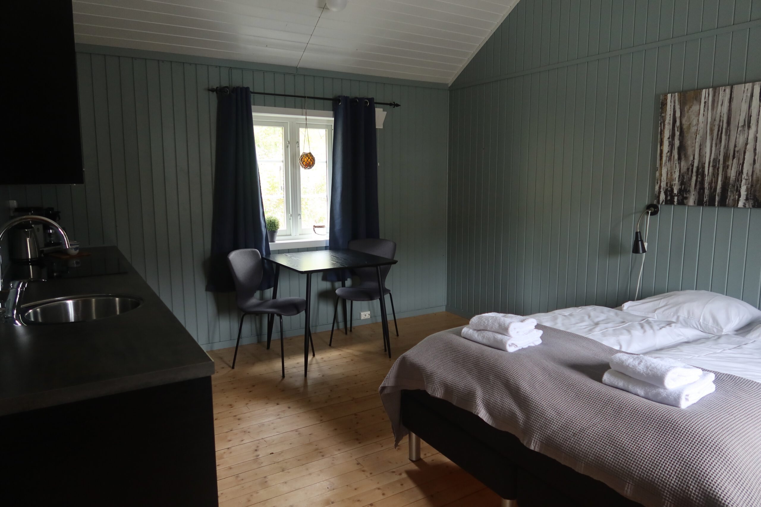 The Tide Fisherman Cabins Sørvågen interior with combined bedroom and kitchenette, double bed with towels, dining table by the window and wooden floors in a cozy rorbu cabin