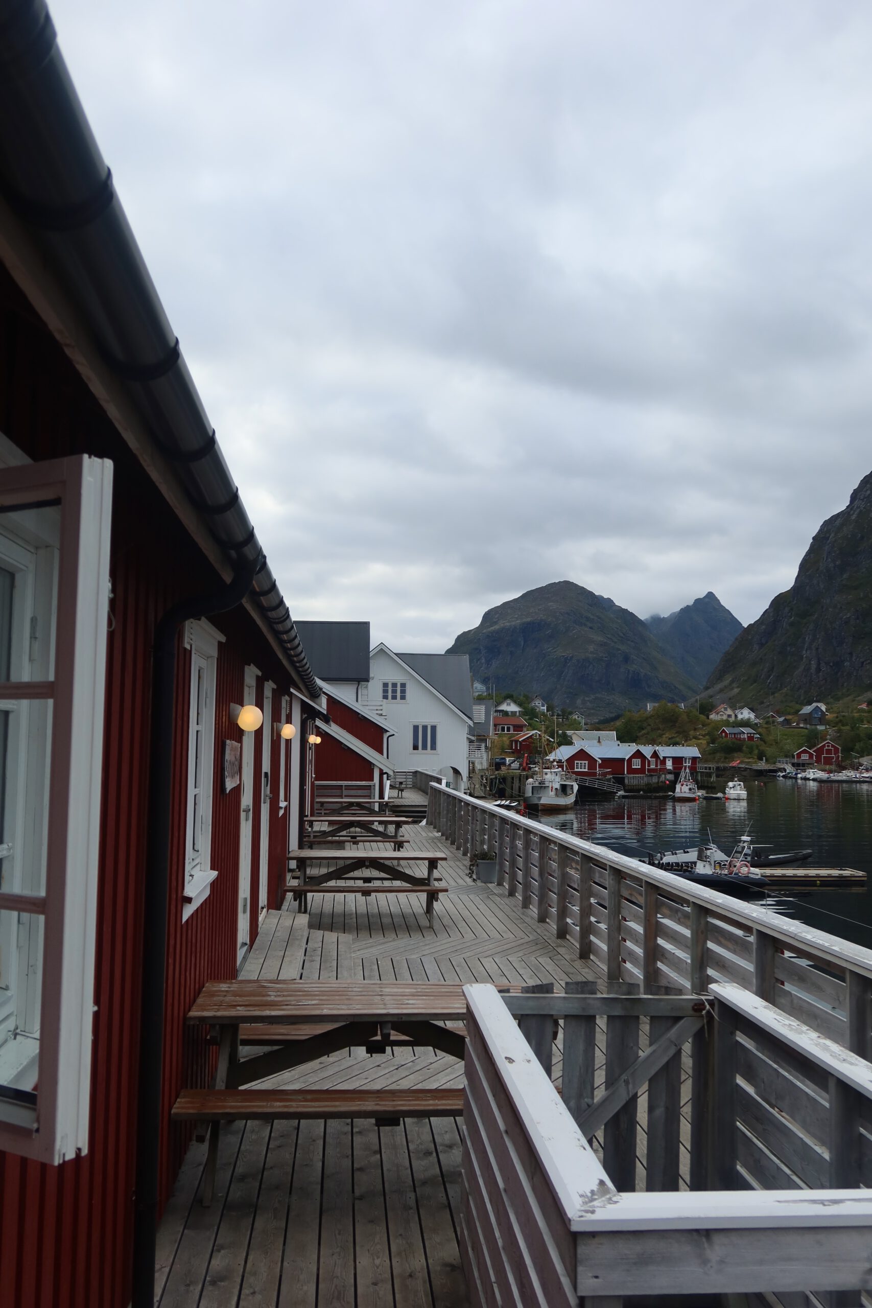 Wooden boardwalk and outdoor seating area at The Tide Fisherman Cabins Sørvågen with red fisherman cabins, harbor reflections and surrounding Lofoten mountains