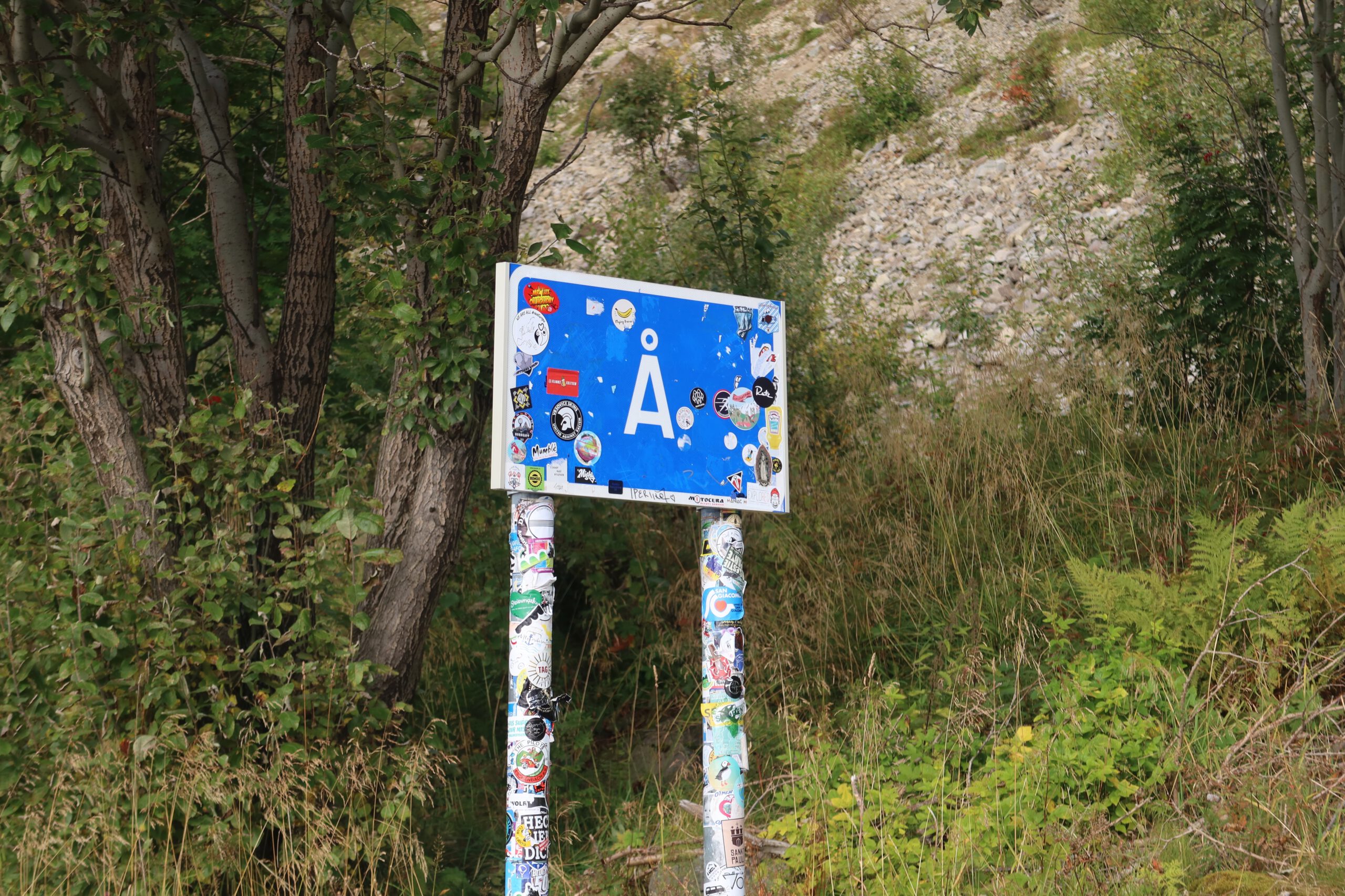 Blue Å village sign covered with travel stickers near Sallybua Lofoten restaurant surrounded by nature