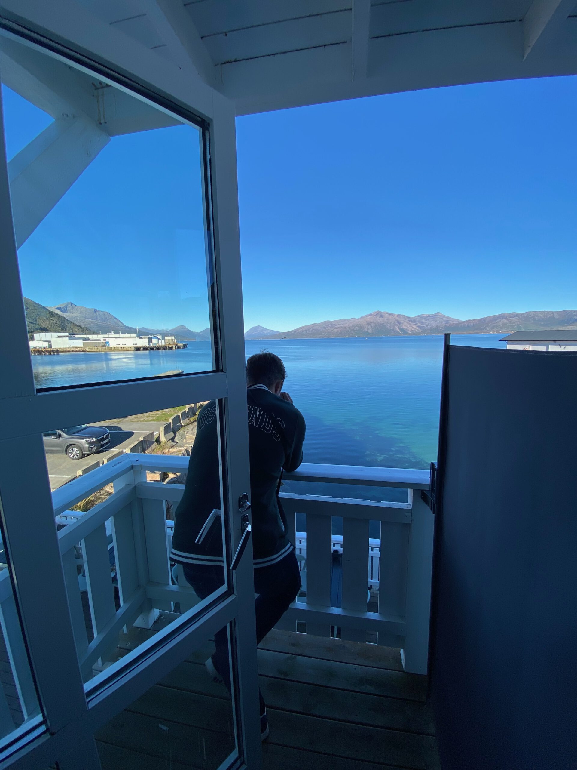 Peter Smit enjoying the balcony view from Lødingen Brygge Hotel Norway room, overlooking the fjord with clear blue water and mountain landscape in Northern Norway