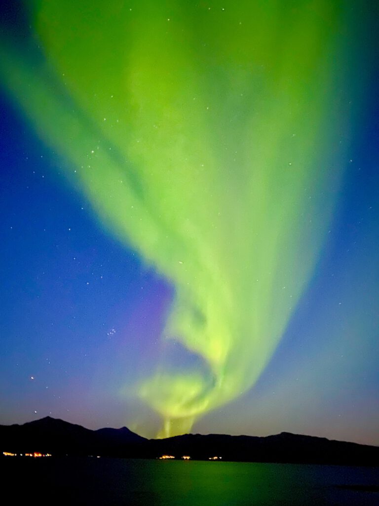 Aurora borealis above Hjertholmen island in Lødingen, northern lights glowing over the harbor and village lights reflected on the water in Northern Norway