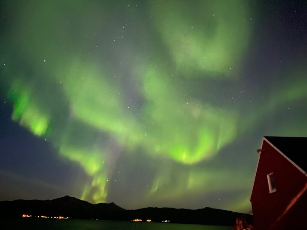 Northern Lights above Lødingen Brygge Hotel Norway, green aurora in the night sky with a wooden building and distant shoreline lights in Northern Norway