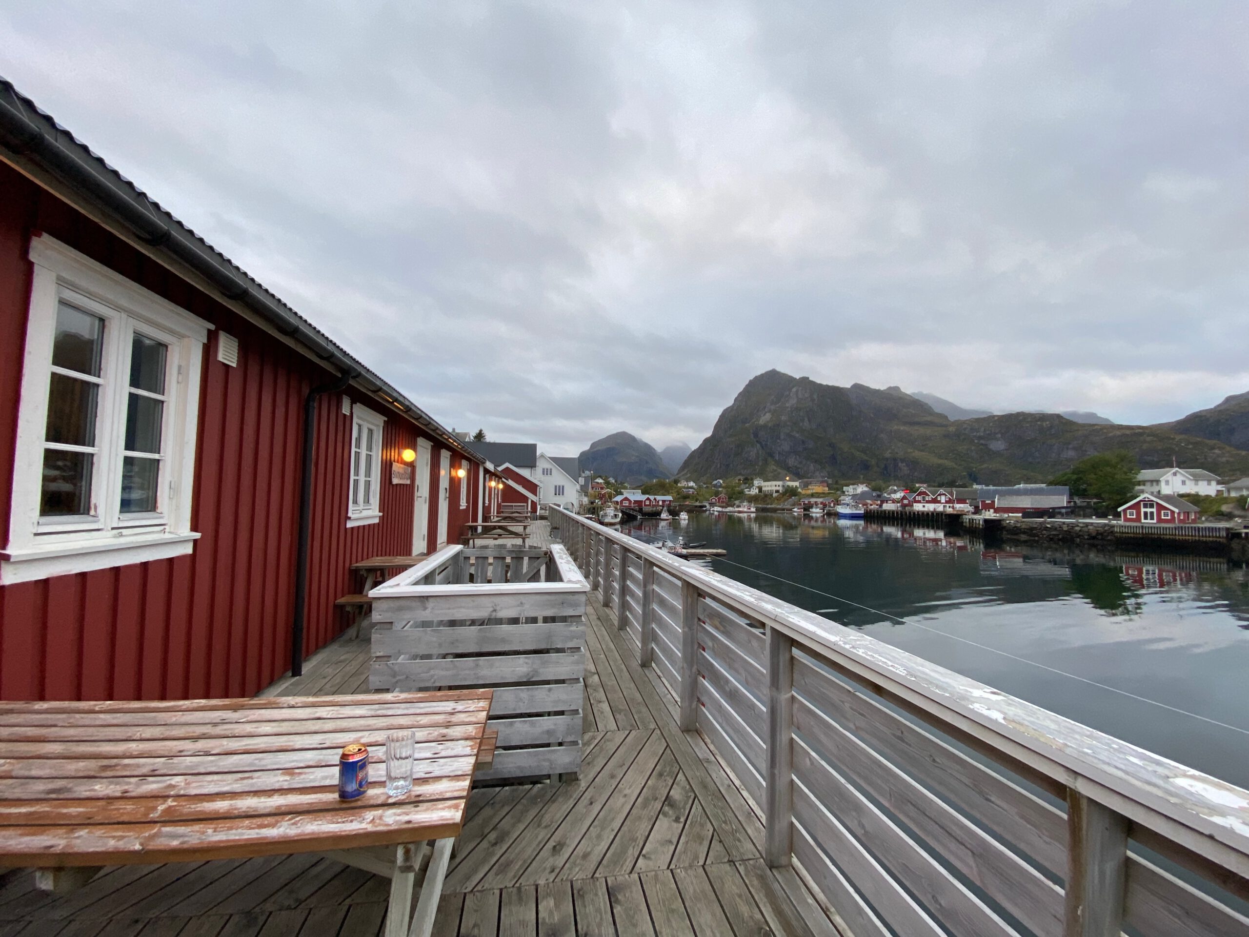 The Tide Fisherman Cabins Sørvågen waterfront view with red rorbuer cabins, wooden terrace, calm harbor water and mountains in the background on the Lofoten Islands