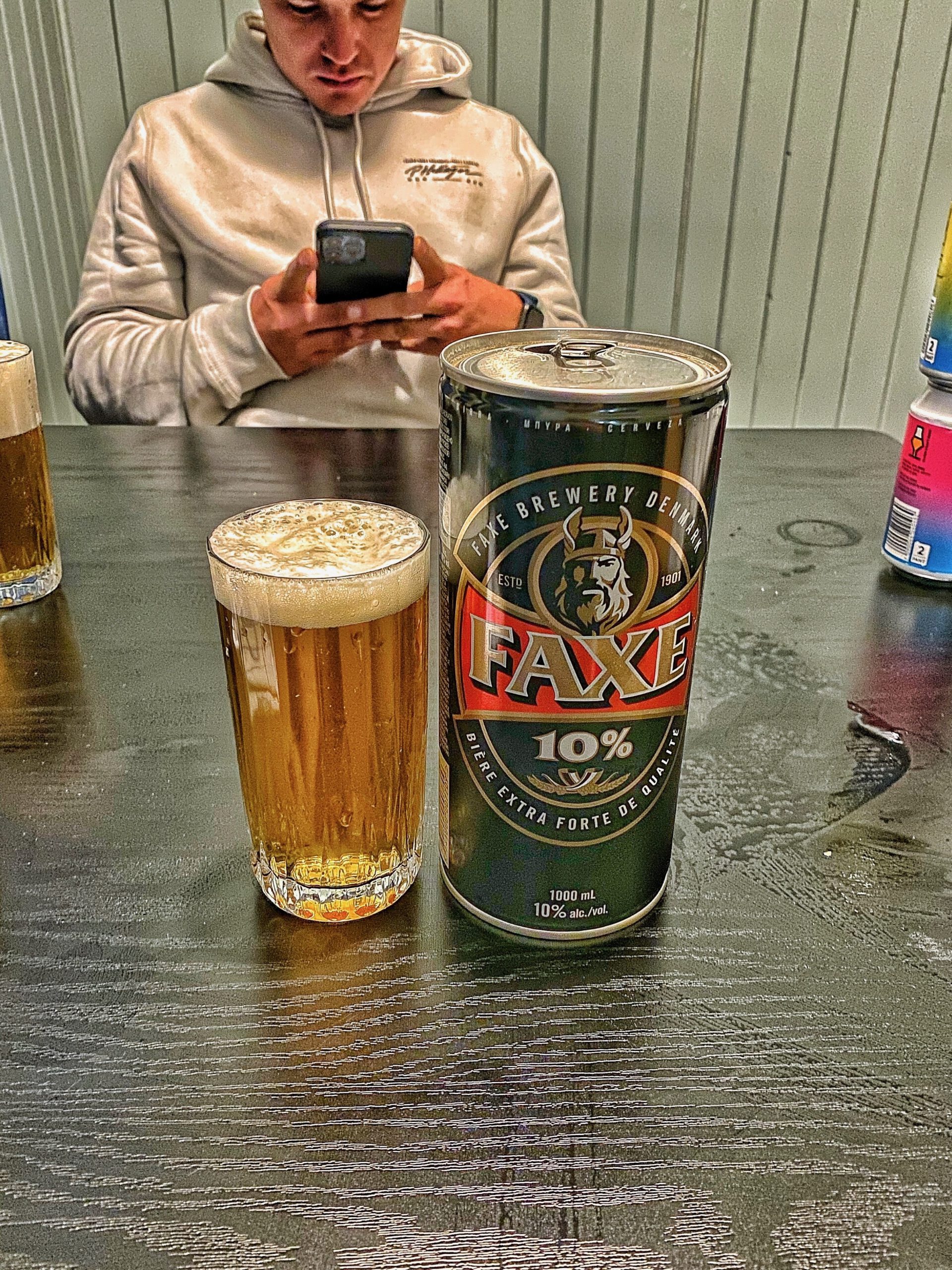 Evening drink moment at The Tide Fisherman Cabins Sørvågen with a glass of beer and a large Faxe strong beer can on the table inside the cozy cabin after a long day in the Lofoten Islands