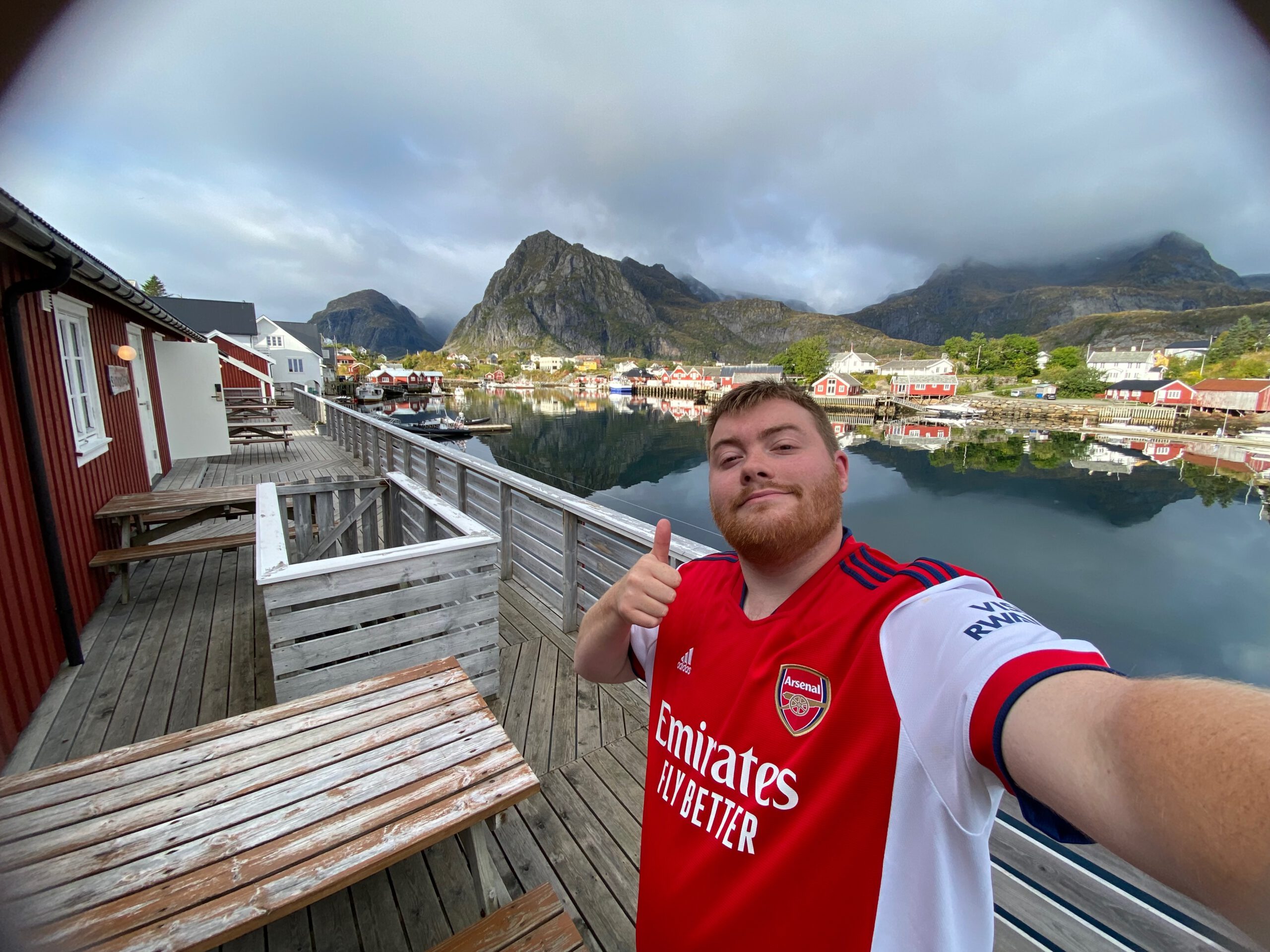 The Tide Fisherman Cabins Sørvågen waterfront boardwalk selfie of Timo van den Berg, Timeuw, with an Arsenal Jersey on, with red rorbuer cabins, calm harbor water and dramatic Lofoten mountains in the background