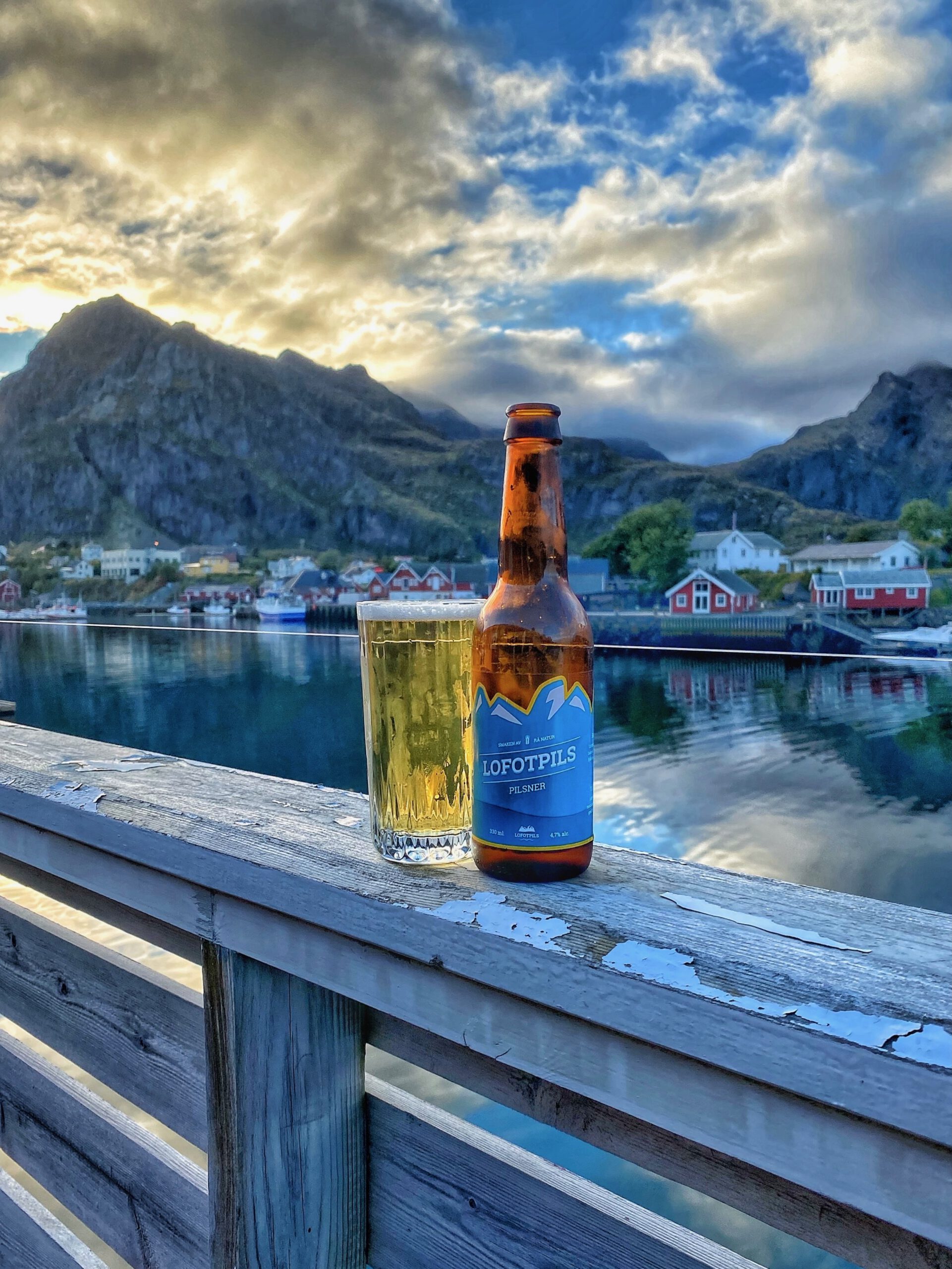 Evening view at The Tide Fisherman Cabins Sørvågen with a glass and bottle of Lofotpils beer on the wooden deck overlooking the calm harbor and dramatic Lofoten mountains at sunset