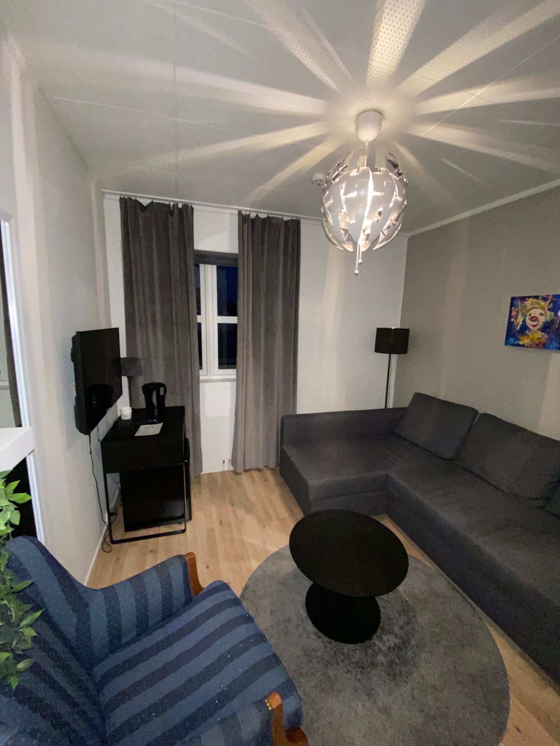 Spacious living room at Mosjøen Romutleie Sentrum 3 featuring a large sofa, armchair, round coffee table, wooden floor, window with curtains, and modern ceiling lamp, a practical budget accommodation interior in Mosjøen, Norway